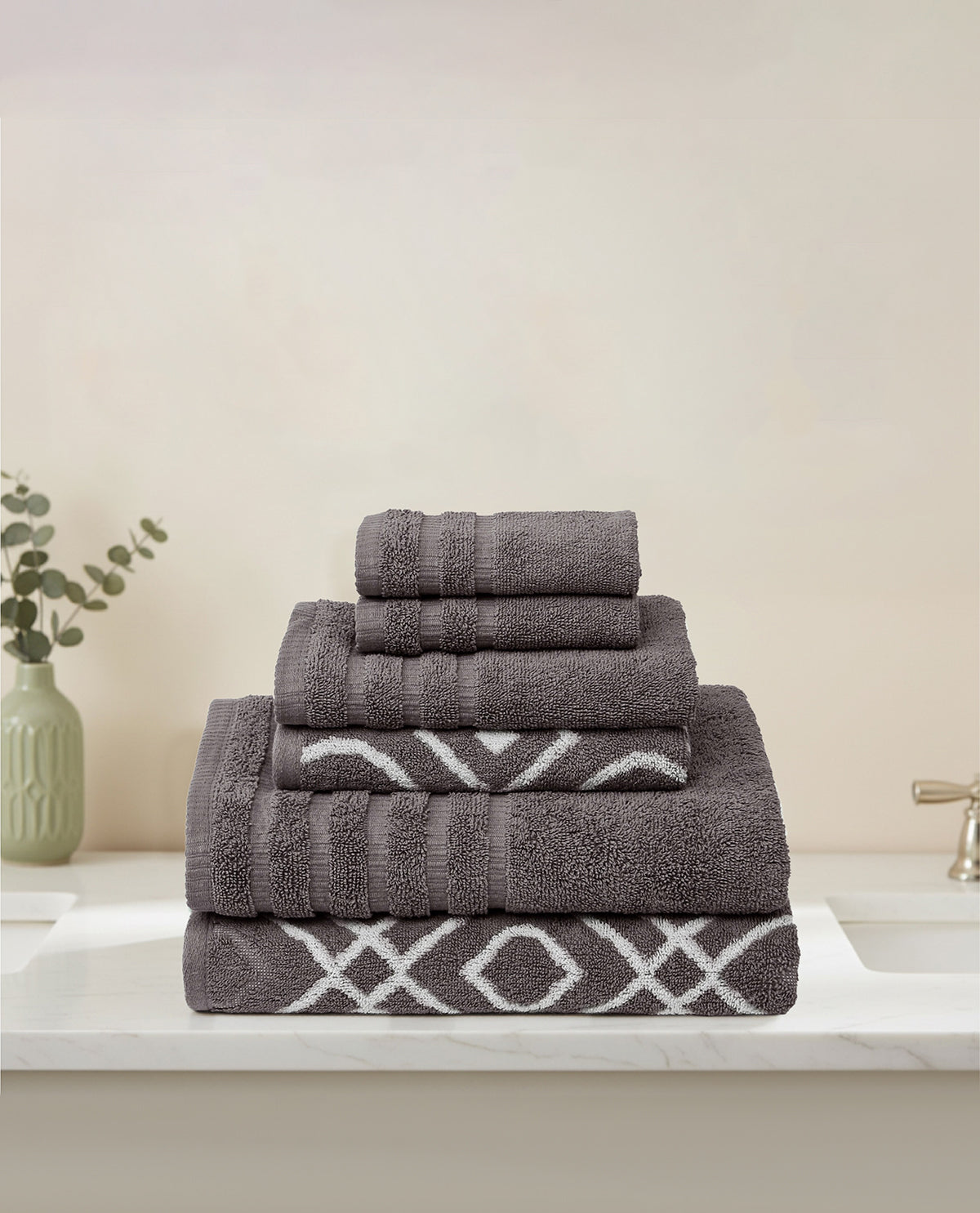 A stack of two folded gray bath towels, 2 hand towels, and two washcloths with white geometric patterns, displayed on a white marble countertop in a bathroom setting.
