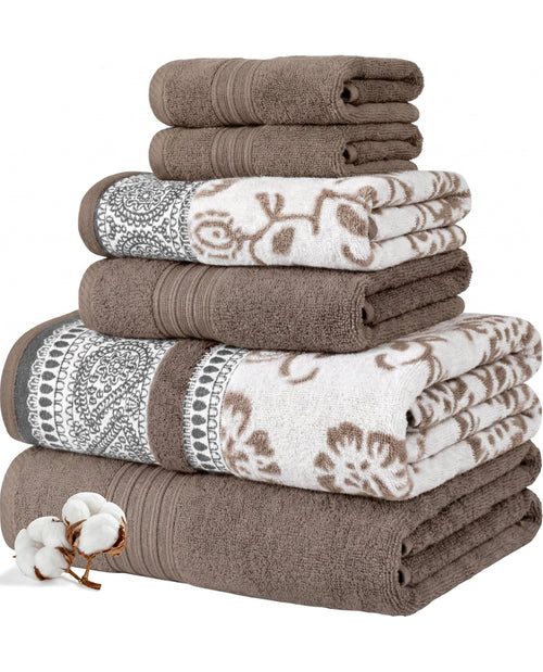 A stack of folded bath towels in brown and white with floral and paisley patterns, shown on a white background with a small cotton boll sprig in the lower left corner.