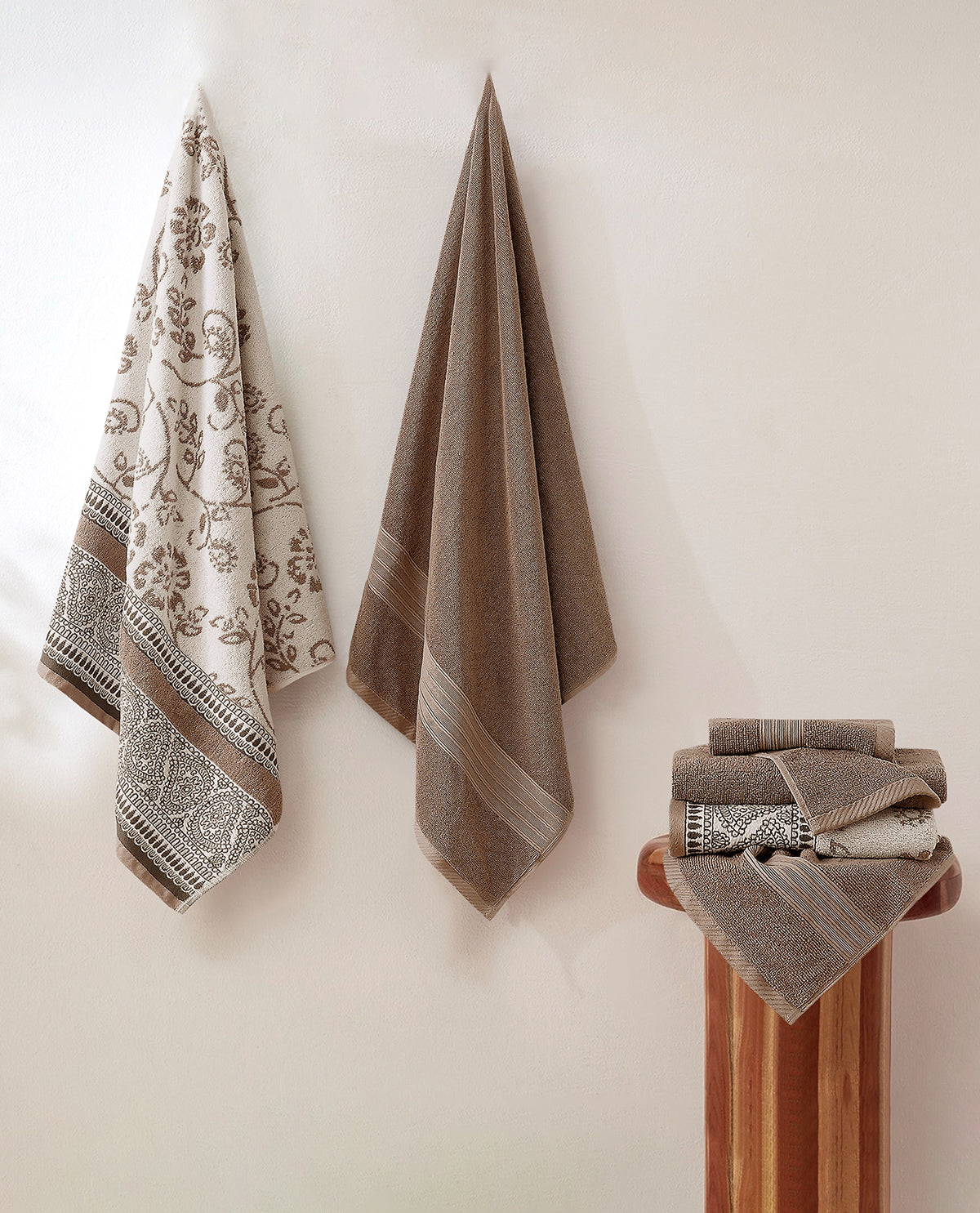 A set of beige and brown towels, including a patterned bath towel hanging on the left, a plain brown towel in the center, and a folded stack of towels on a wooden stand on the right, all set against a warm off-white wall.