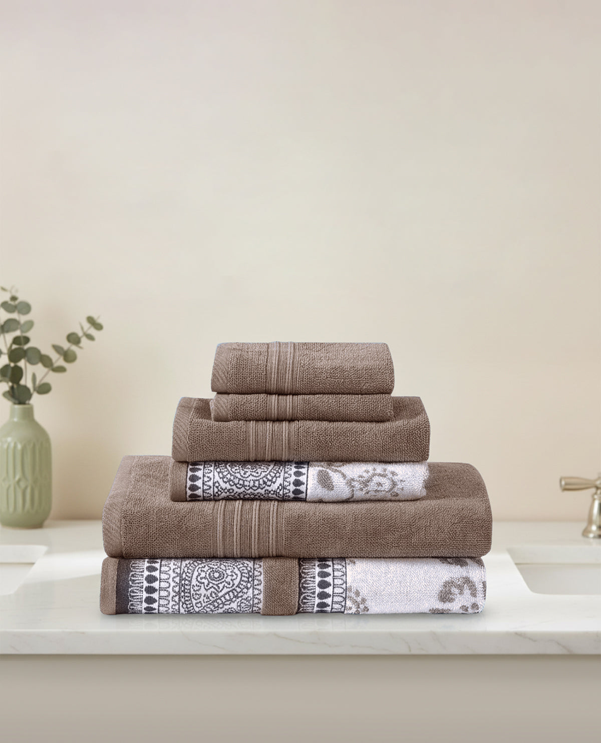 A stack of two folded bath towels, two hand towels, and two washcloths in a taupe-brown color, with two towels featuring a black and white geometric patterned border, displayed on a white marble countertop next to a sink and a small vase with greenery.