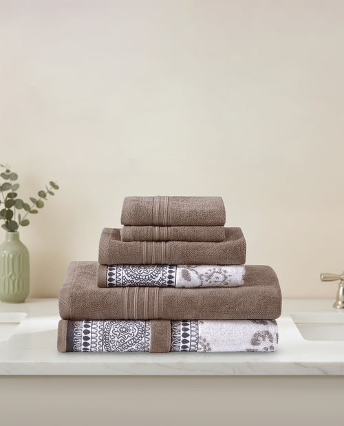 A stack of two folded bath towels, two hand towels, and two washcloths in a taupe-brown color, with two towels featuring a black and white geometric patterned border, displayed on a white marble countertop next to a sink and a small vase with greenery.
