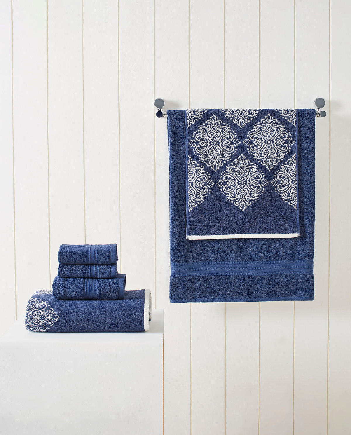 A set of navy blue towels with white damask patterns, shown hanging and folded on a white wood-paneled wall.