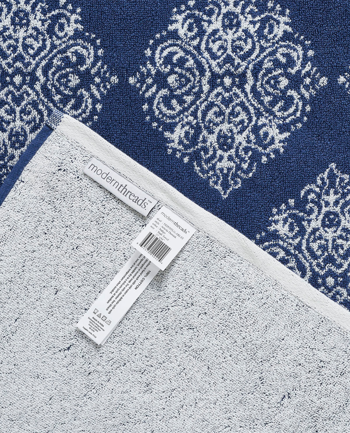 Close-up of a folded bath towel showing a navy blue and white damask pattern on the top and a light gray textured backing on the bottom, with a visible Modern Threads brand tag.