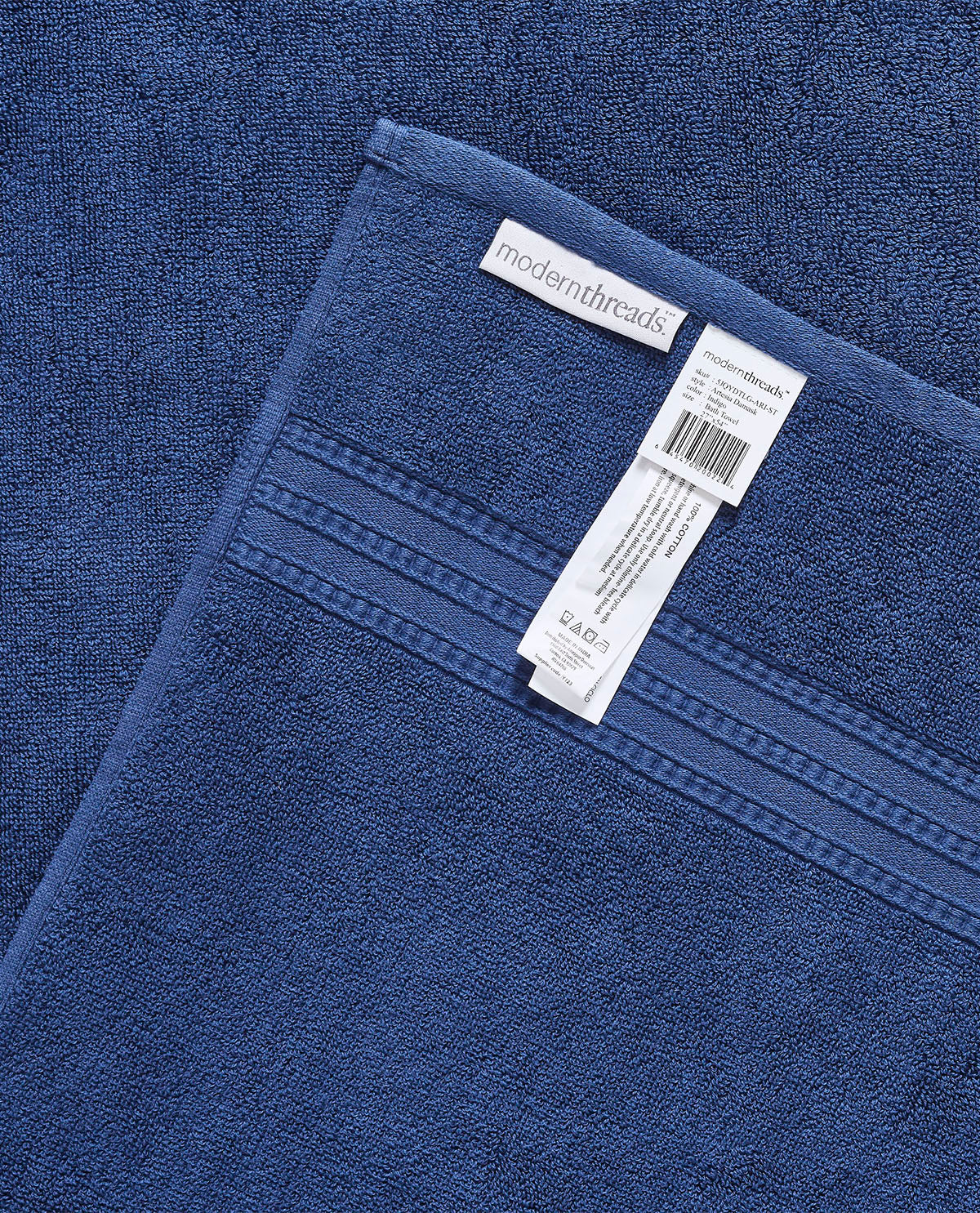 A close-up of a folded blue towel with a white sewn-in label reading 'modernthreads' and a hanging product tag, showing the terry texture and stitched border detail.