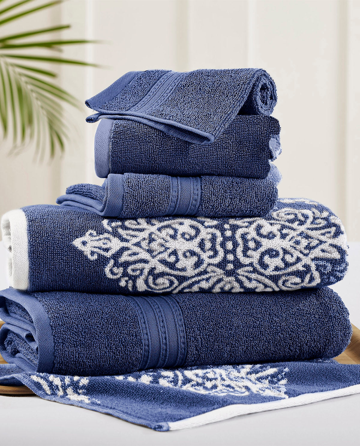 A stack of navy blue and white patterned cotton towels, including hand towels and bath towels, arranged on a light surface with a soft neutral background and a hint of green leaves in the corner.
