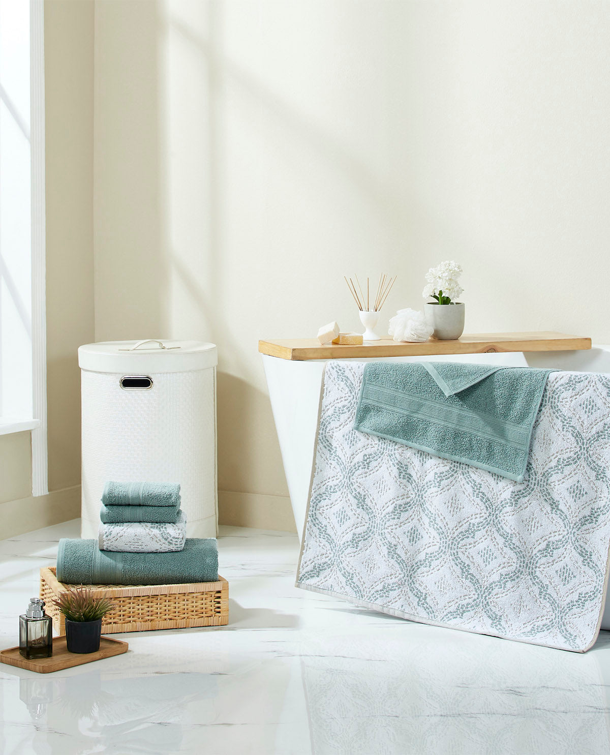 A styled bathroom scene with folded towels and bath accessories arranged around a freestanding bathtub. The most prominent sellable item appears to be the large patterned bath mat or towel cover in white and pale blue-gray, with matching folded towels nearby and a white laundry hamper.