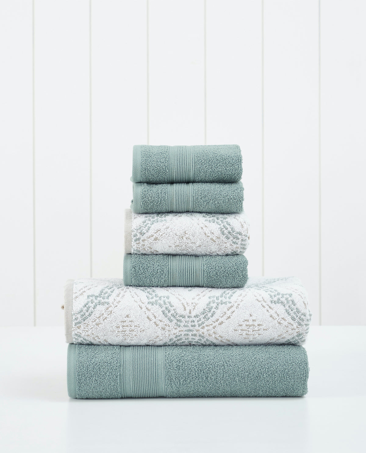 A stack of six folded bath towels in a soft sage green and white patterned set, arranged neatly on a white surface against a white paneled background.