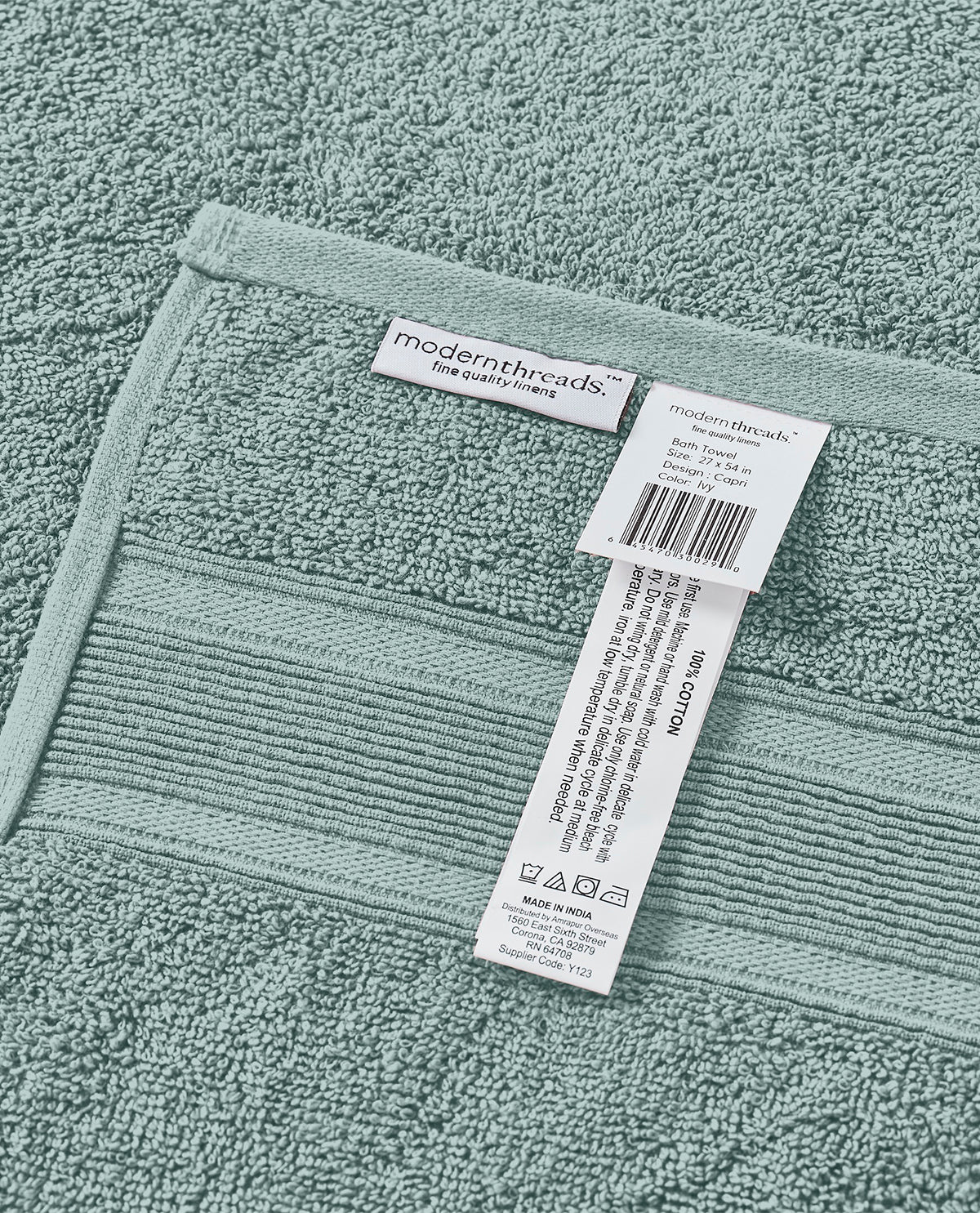 A close-up of a sage green cotton towel with a ribbed border and two white paper tags attached, showing the brand 'modernthreads'.