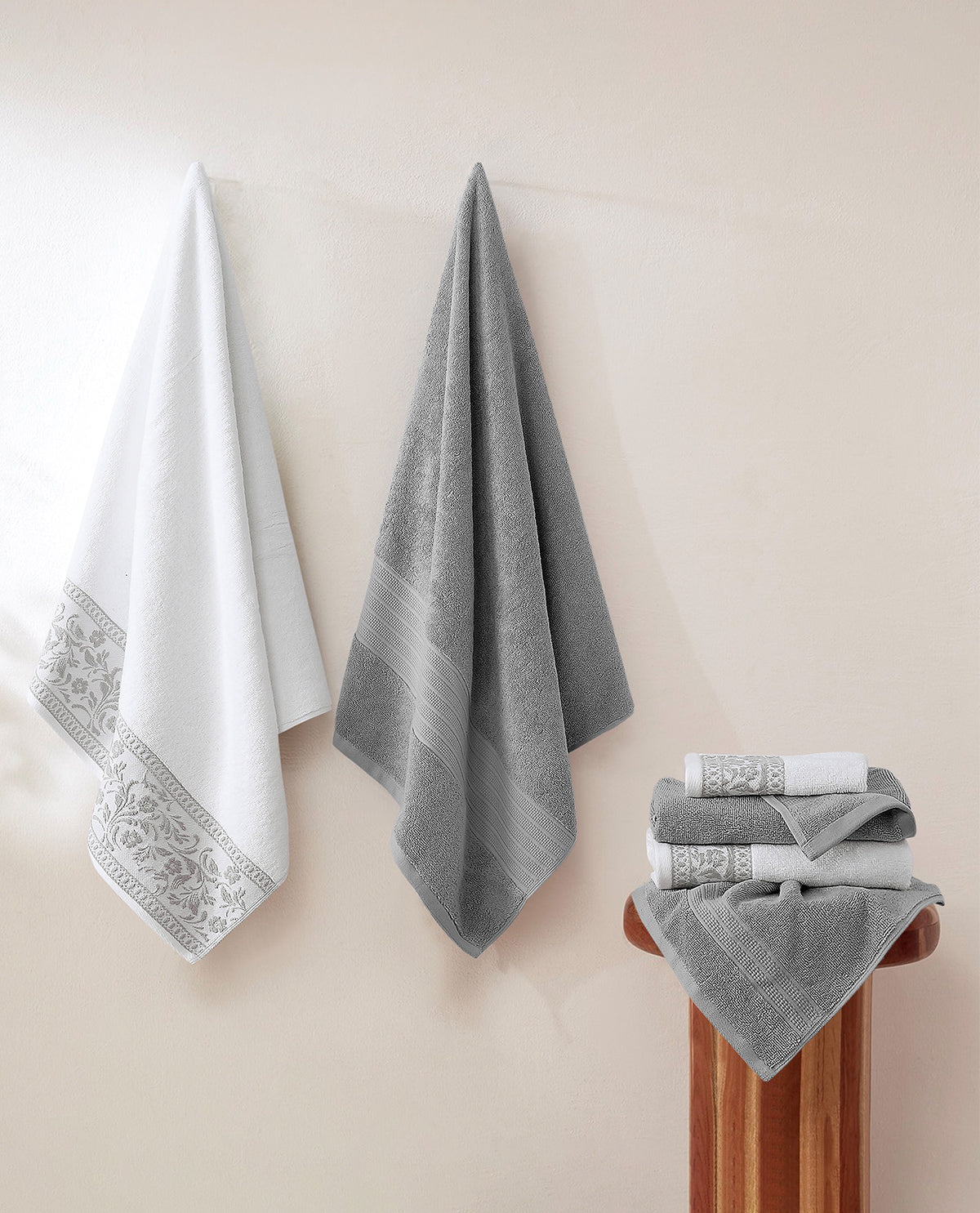 Two bath towels and a folded stack of matching towels with ornate white borders hang on a beige wall, with a wooden stand holding the folded stack.