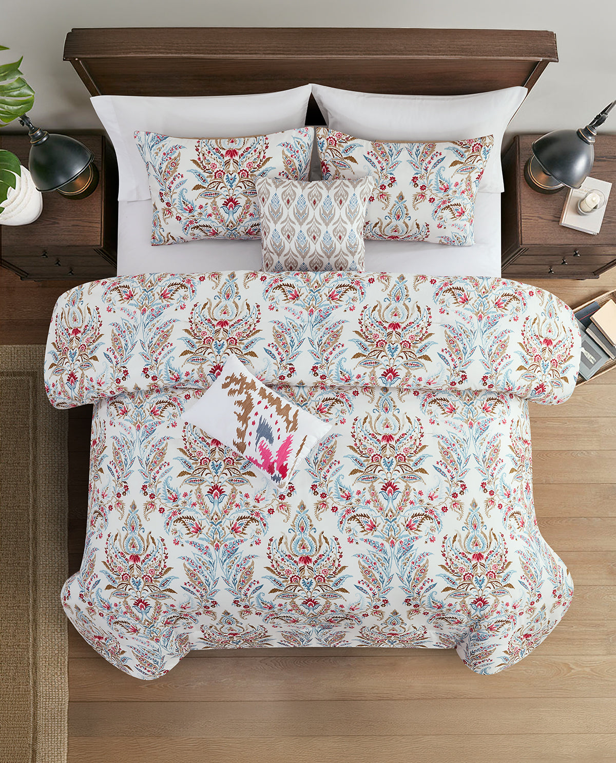 A top-down view of a bed styled with a white duvet cover and matching pillow shams featuring a detailed multicolor paisley and floral pattern in red, blue, and brown. A small decorative pillow with a pink and brown pattern rests on the duvet.