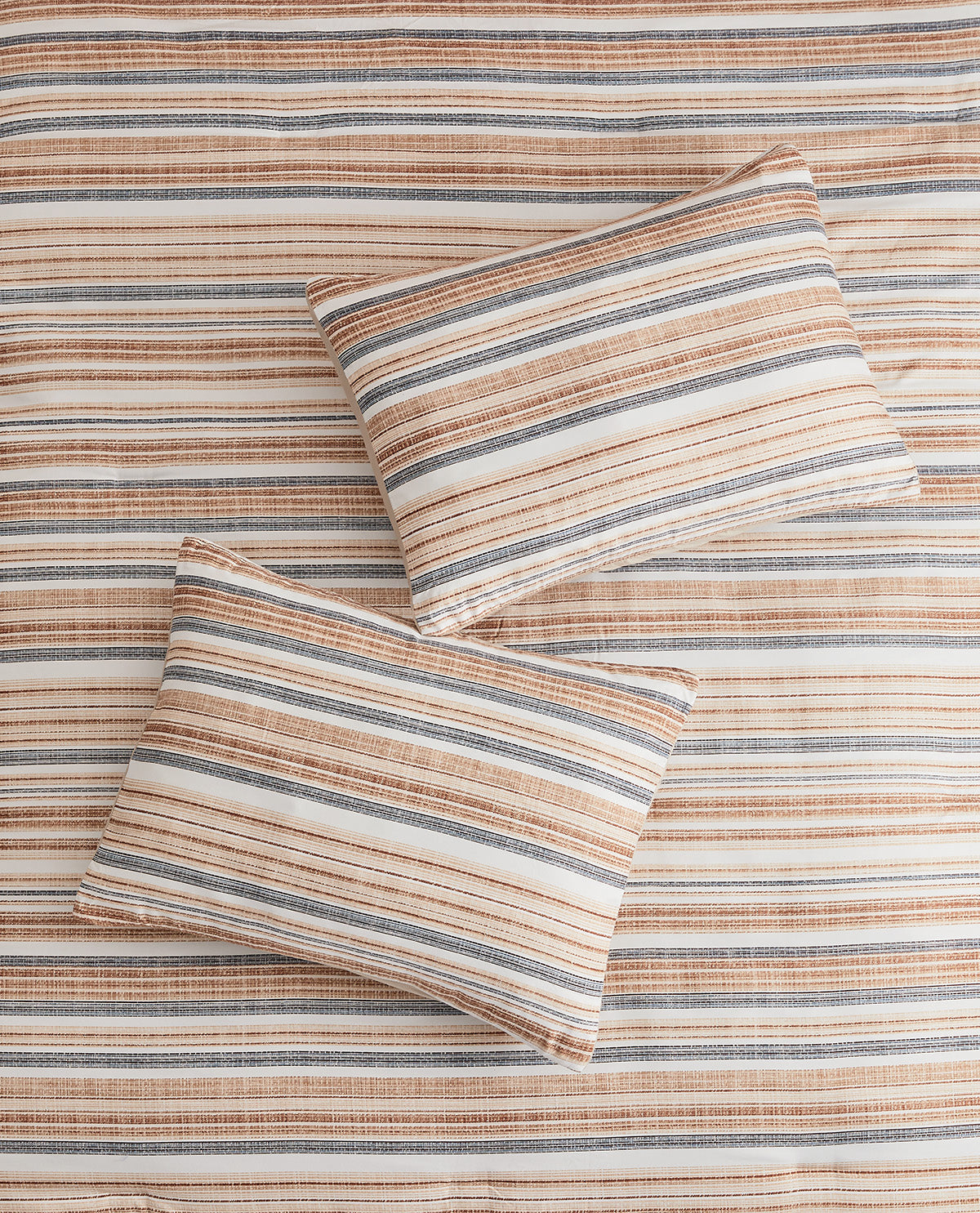 Two striped throw pillows on a matching striped bedspread or blanket in warm beige, cream, brown, and muted gray tones.