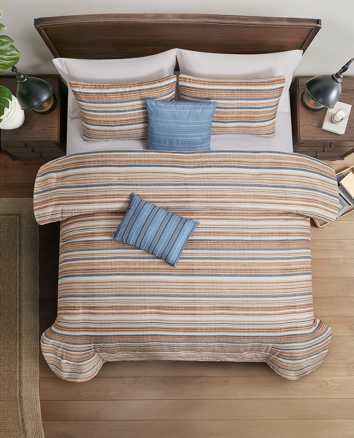 A top-down view of a bed styled with a striped duvet cover and matching pillow shams, featuring two solid blue accent pillows. The bed is set against a dark wood headboard, flanked by matching wooden nightstands with black lamps, on a light wood floor.