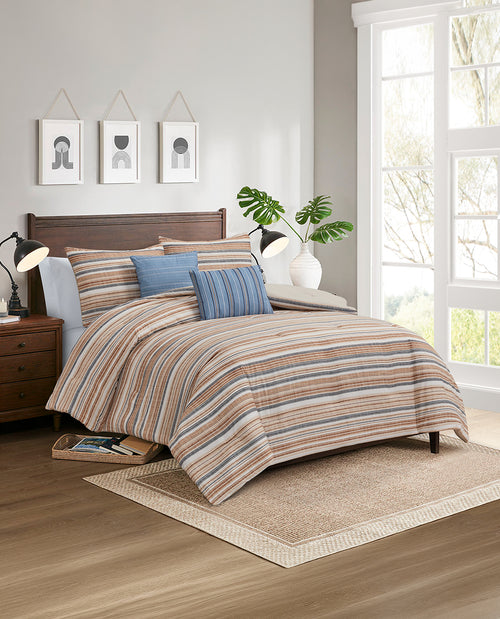A bed dressed in a striped comforter and matching pillow shams in beige, brown, blue, and gray tones, styled in a bright bedroom with wood furniture, lamps, wall art, and a window.
