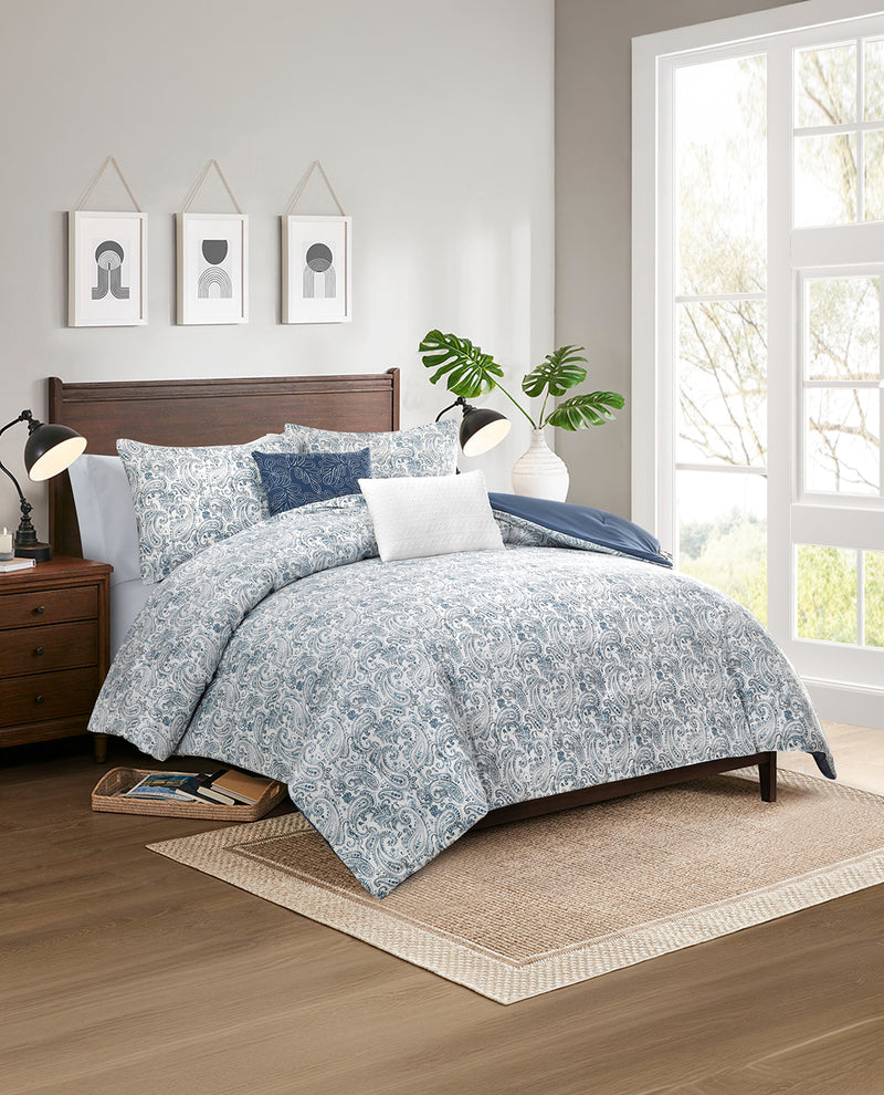 A bed dressed in a light blue and white floral patterned comforter set with matching pillows, styled in a bright bedroom with a wooden headboard, nightstand, window, and rug.
