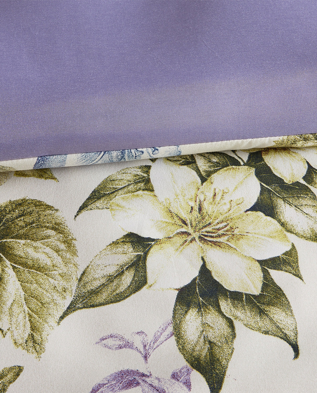 A close-up of a floral fabric with a large cream flower and olive-green leaves on a light background, partially folded over to reveal a solid lavender underside.