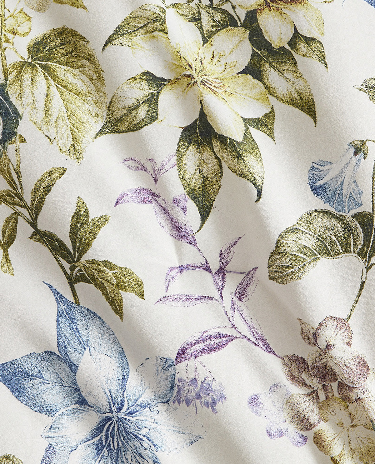 A close-up of a fabric surface featuring a botanical floral print with cream, blue, purple, and olive-green leaves and flowers on an off-white background.
