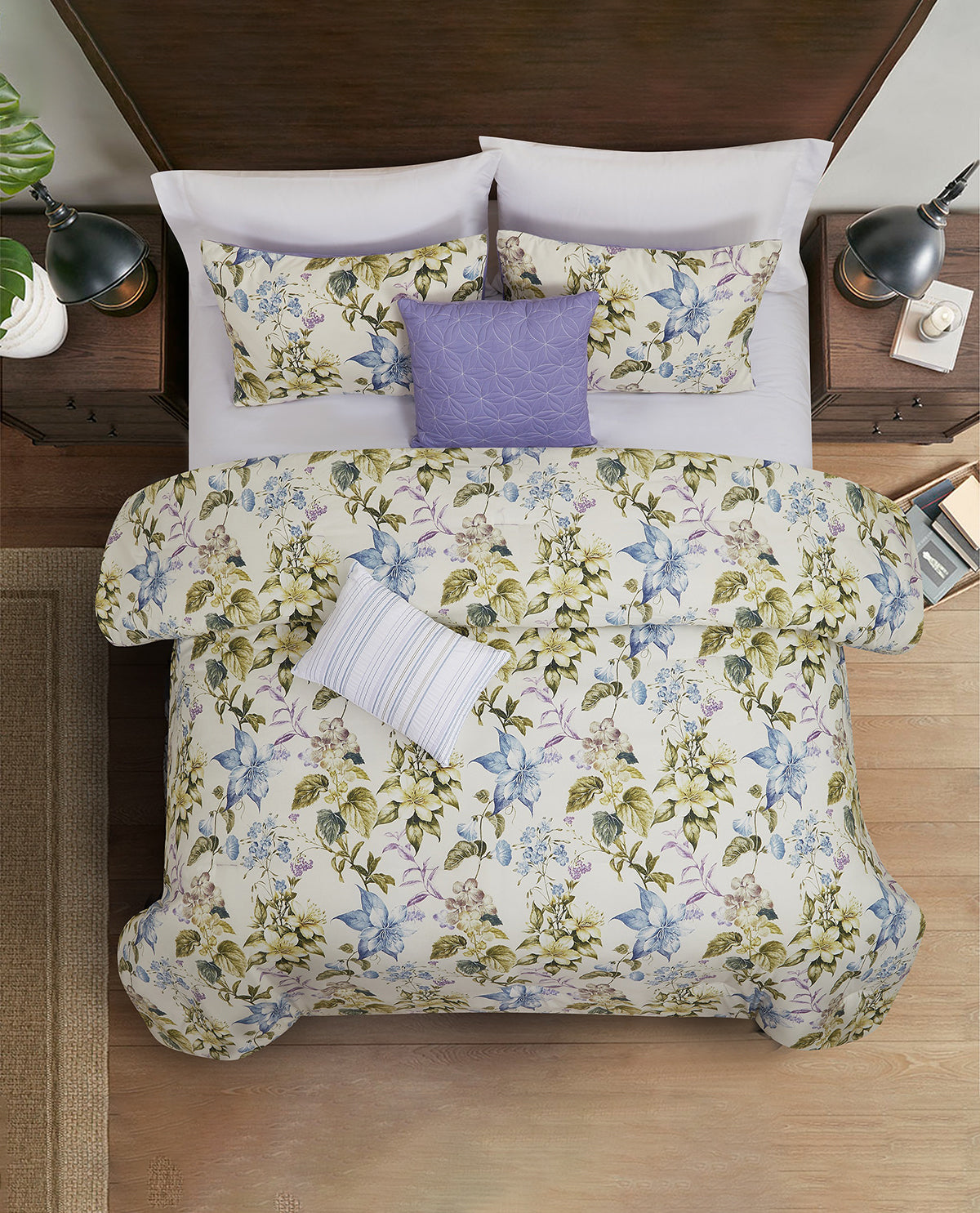 A top-down view of a bed styled with a floral comforter and matching pillow shams, featuring a lavender accent pillow. The bedding has a cream background with blue, green, and purple botanical patterns, set in a modern bedroom with dark wood nightstands and lamps.