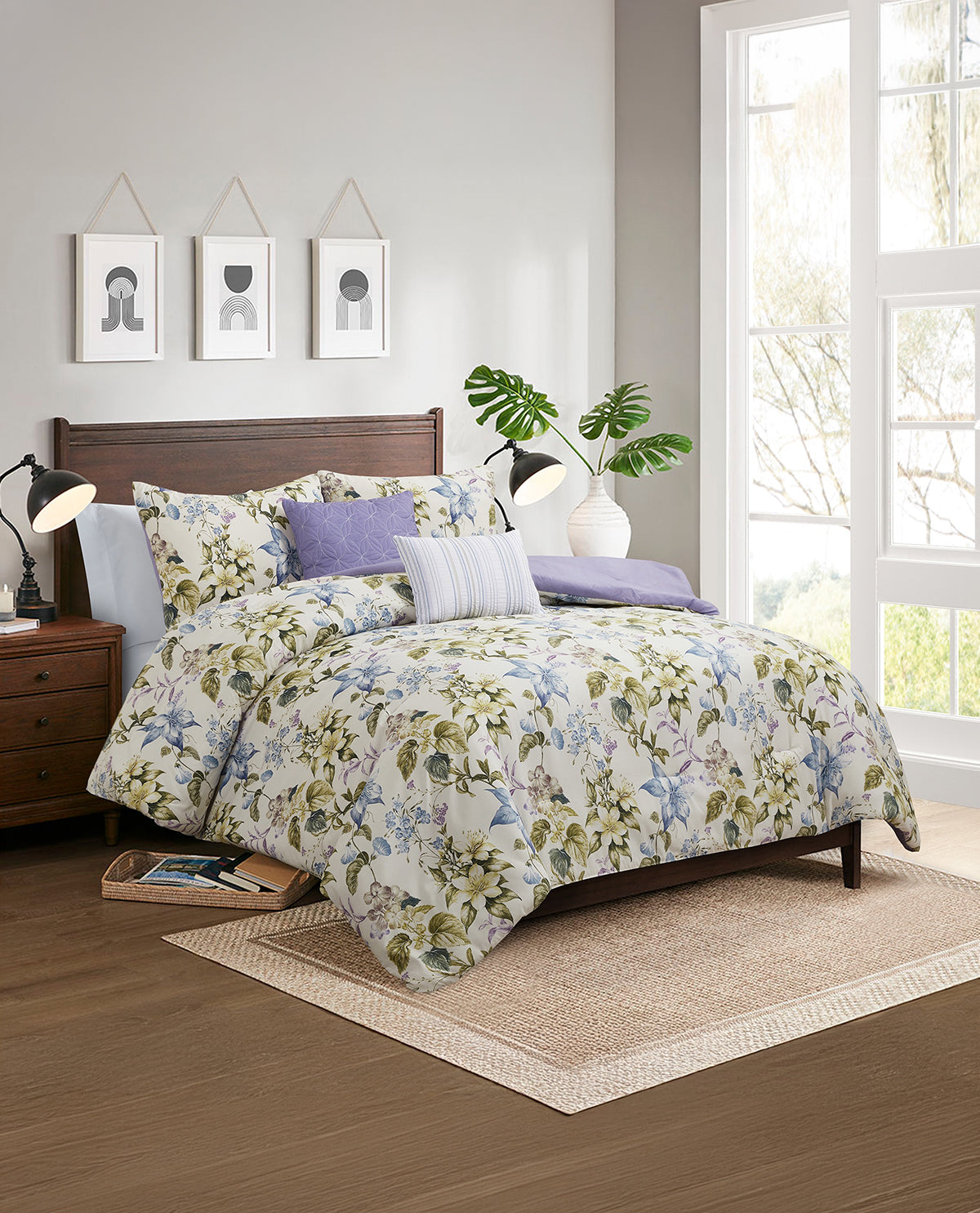 A floral bedding set with a white background, green and blue botanical print, and purple accent pillows, styled on a dark wood bed frame in a bright, modern bedroom.