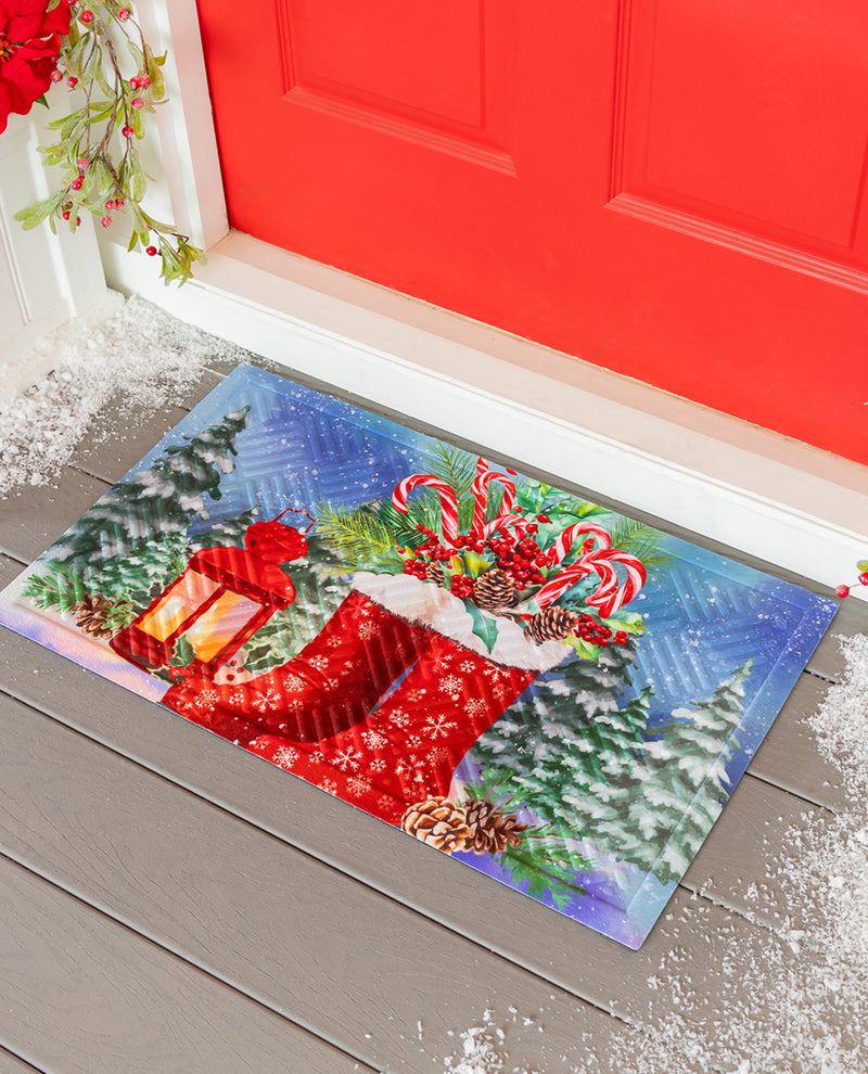A rectangular Christmas-themed doormat placed on a gray porch floor in front of a red door. The mat features a festive winter scene with a red stocking, candy canes, pine trees, a lantern, and snow in the background.