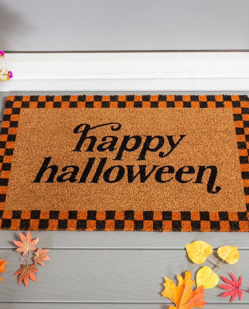 A rectangular coir doormat with 'happy halloween' in black script, bordered by an orange and black checkered pattern, displayed on a gray wooden surface with autumn leaves.