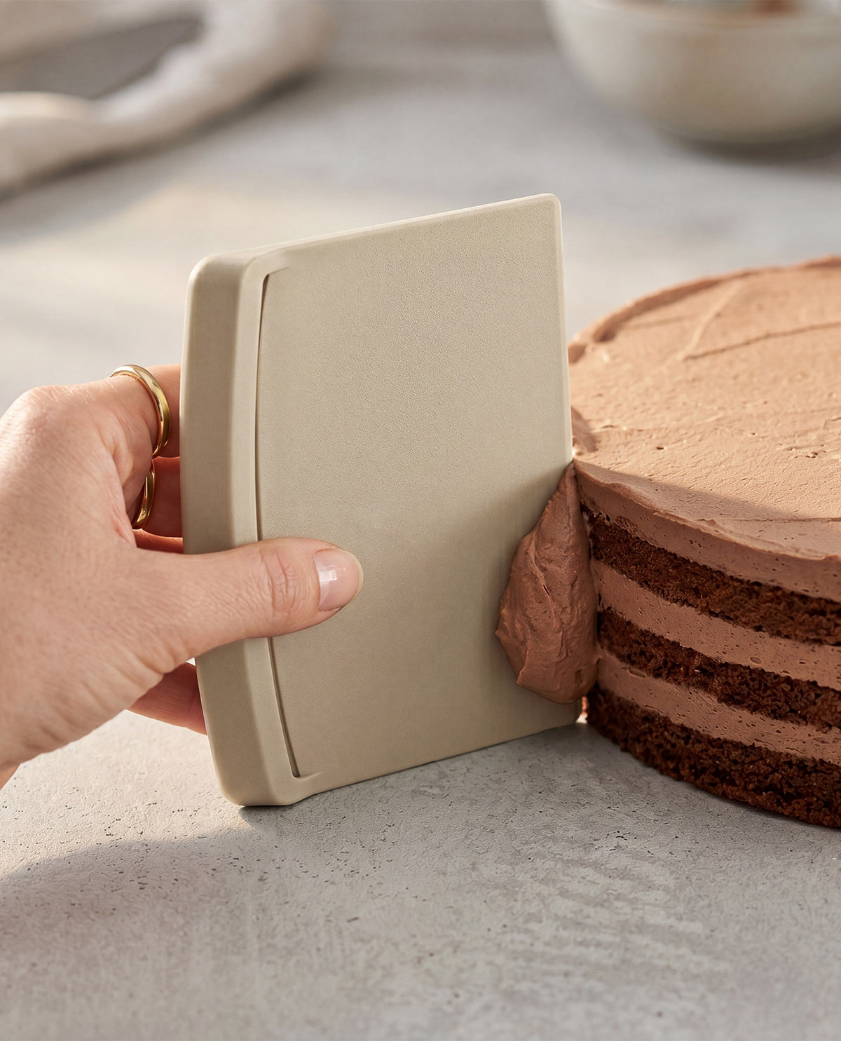 A hand holds a beige rectangular cake scraper against a chocolate cake with smooth chocolate frosting on a light gray surface.