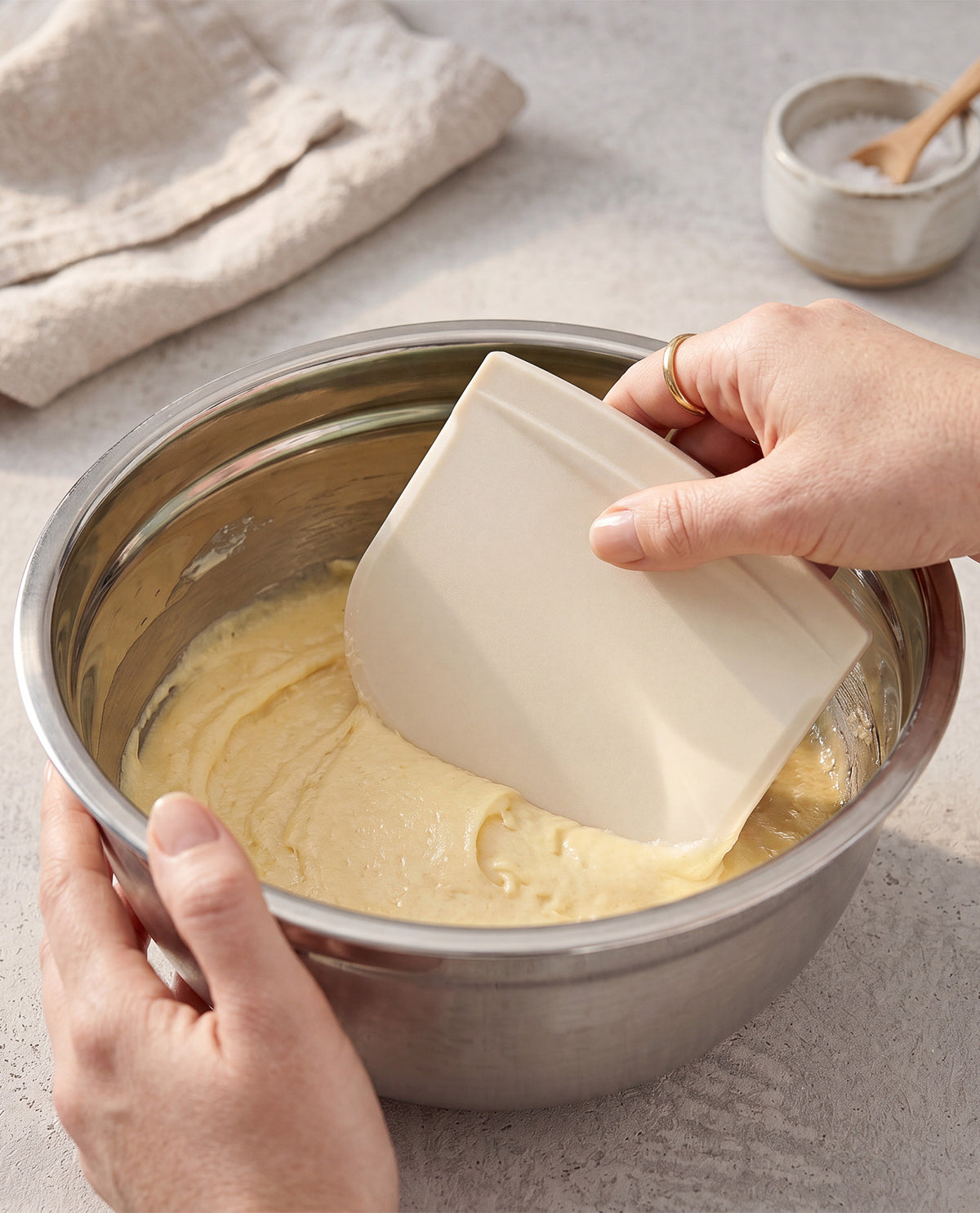 A stainless steel mixing bowl containing pale yellow batter, with a hand holding a white silicone scraper and another hand holding the bowl. A beige cloth and a small dish with a wooden spoon are blurred in the background on a light stone surface.