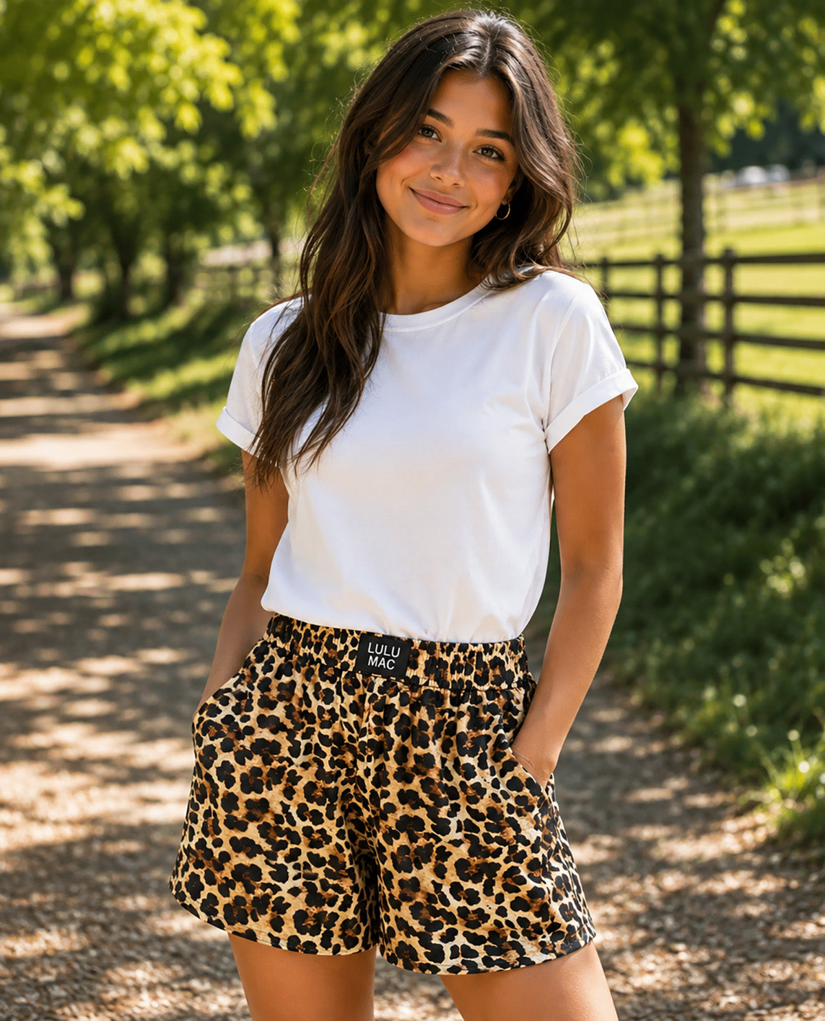 A woman standing on a sunlit country path wearing a white short-sleeve T-shirt and leopard-print shorts with a black belt. Trees, grass, and a wooden fence are visible in the background.