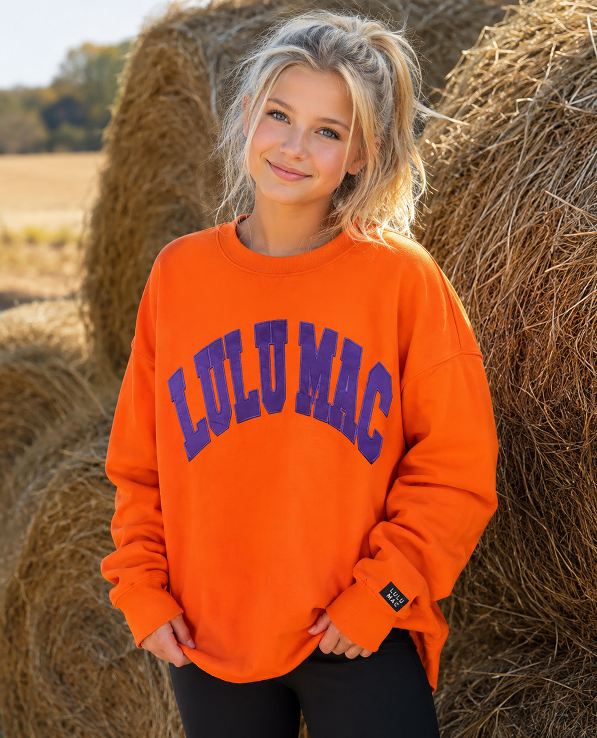 A young woman with blonde hair in a ponytail stands outdoors in front of hay bales, wearing an oversized bright orange sweatshirt with 'LULUmac' printed in dark blue arched letters across the chest.