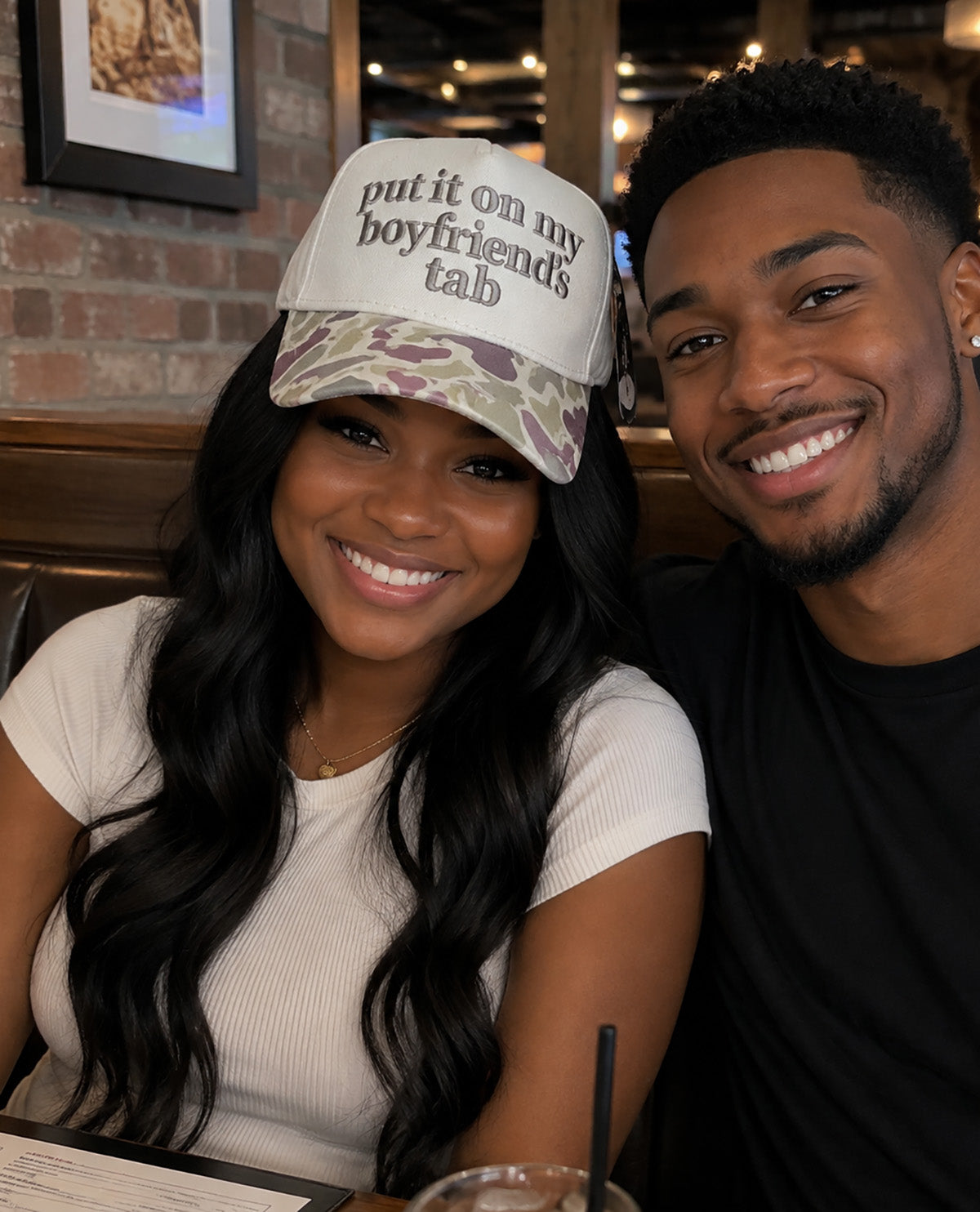 A smiling woman wearing a white trucker hat with the text 'put it on my boyfriend's tab' and a camouflage pattern brim, seated next to a smiling man in a restaurant setting.