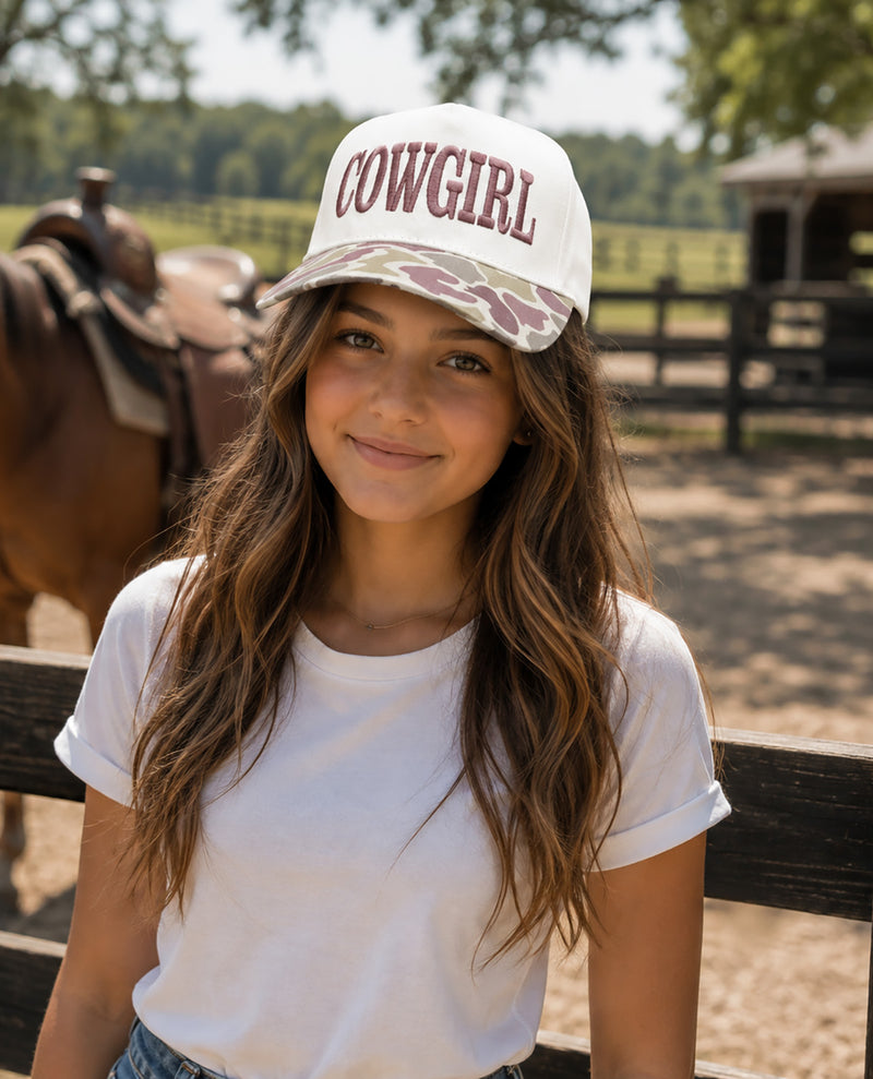 A young woman smiling outdoors in a white trucker-style baseball cap with 'COWGIRL' embroidered on the front and a camouflage brim, wearing a white t-shirt and sitting behind a wooden fence.
