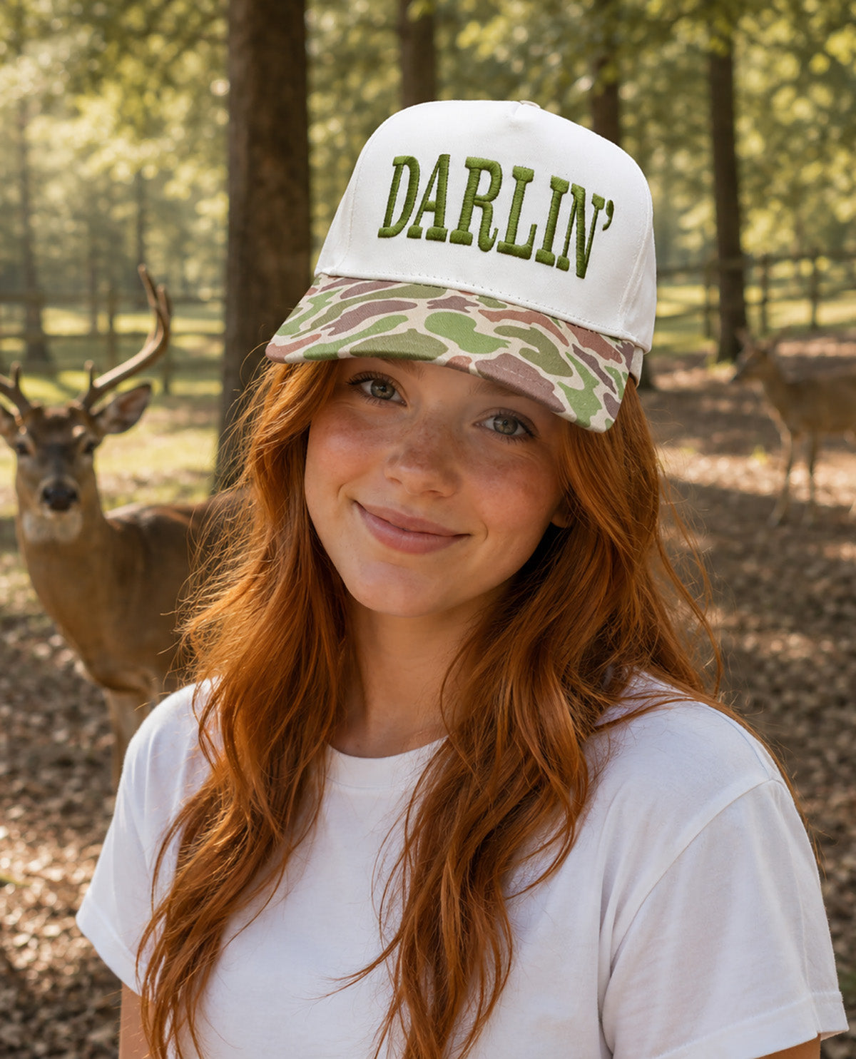 A smiling young woman with long red hair wears a white baseball cap with green 'DARLIN'' lettering and a green-and-brown camouflage brim, standing outdoors in a wooded area with deer in the background.