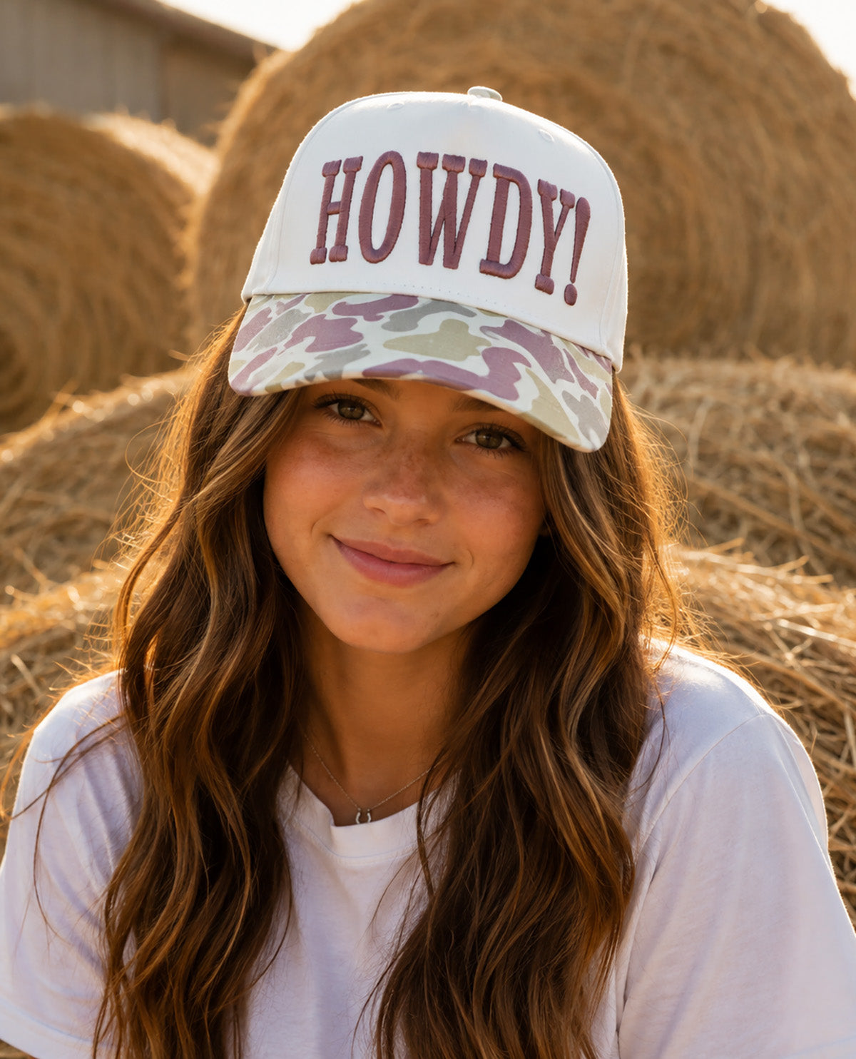 A smiling young woman outdoors in front of hay bales wears a white baseball cap with the word "HOWDY!" in burgundy lettering and a camouflage-patterned visor.