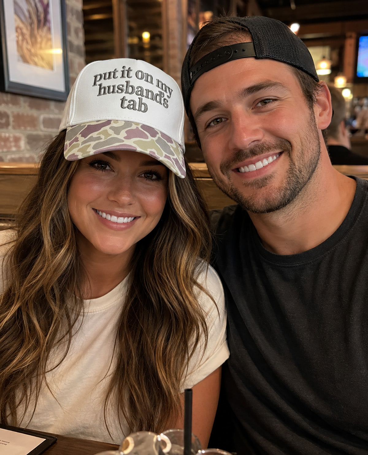 A smiling couple posing together in a restaurant booth. The woman wears a white trucker hat with camouflage brim and the text 'put it on my husband's tab' in black lettering, while the man wears a black baseball cap backwards.