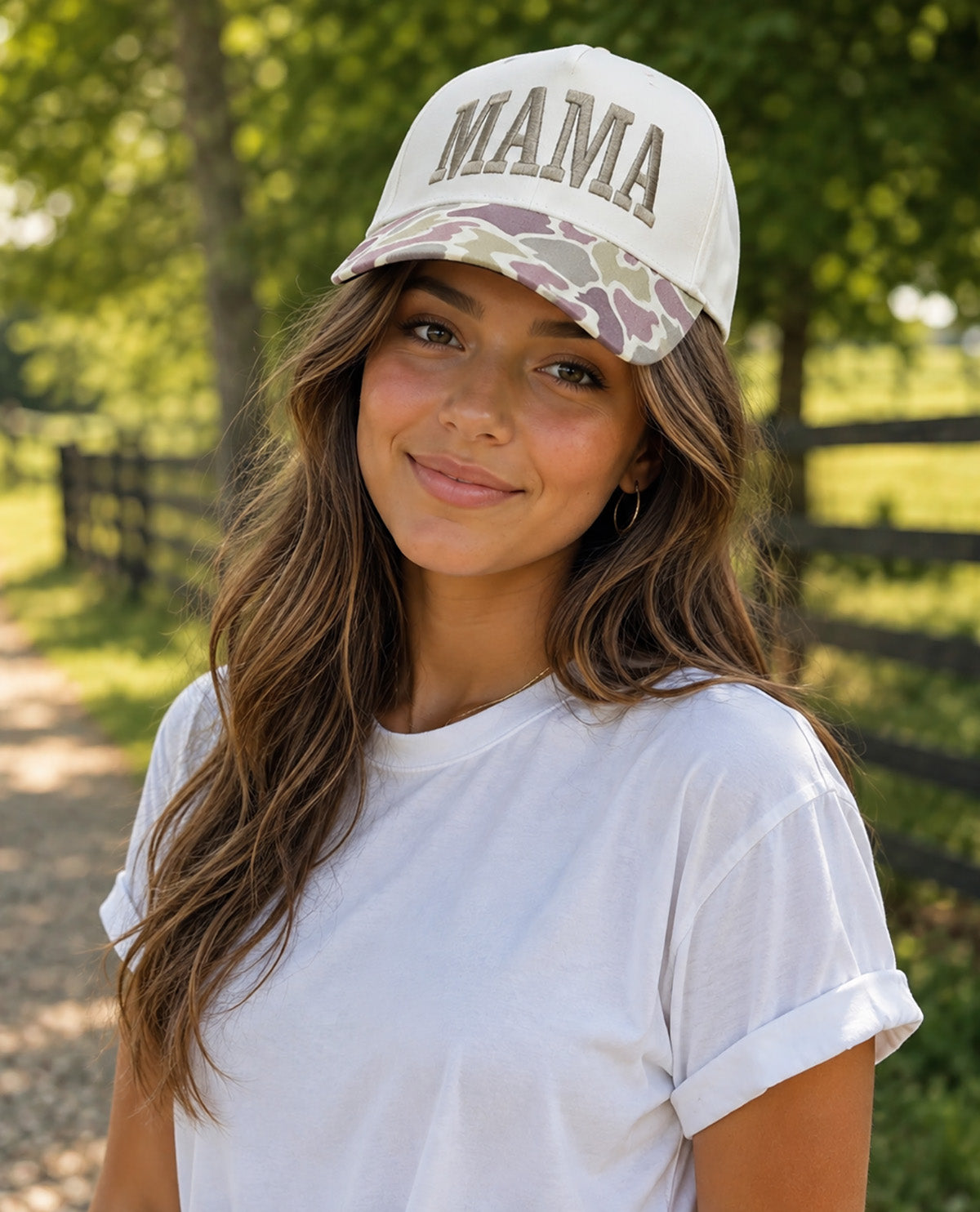 A woman wearing a white baseball cap with 'MAMA' embroidered on the front and a camouflage brim, paired with a white t-shirt, set against a blurred outdoor background of trees and a fence.