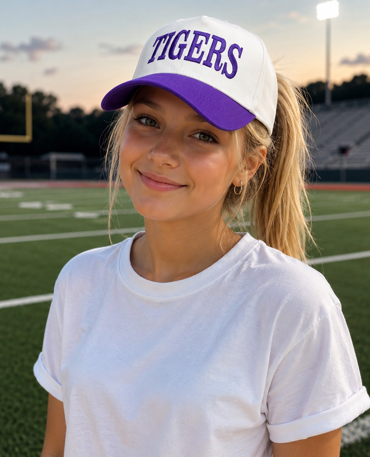 A young woman smiling on a high school football field at sunset, wearing a white and purple baseball cap with 'TIGERS' embroidered in purple on the front, paired with a plain white t-shirt.