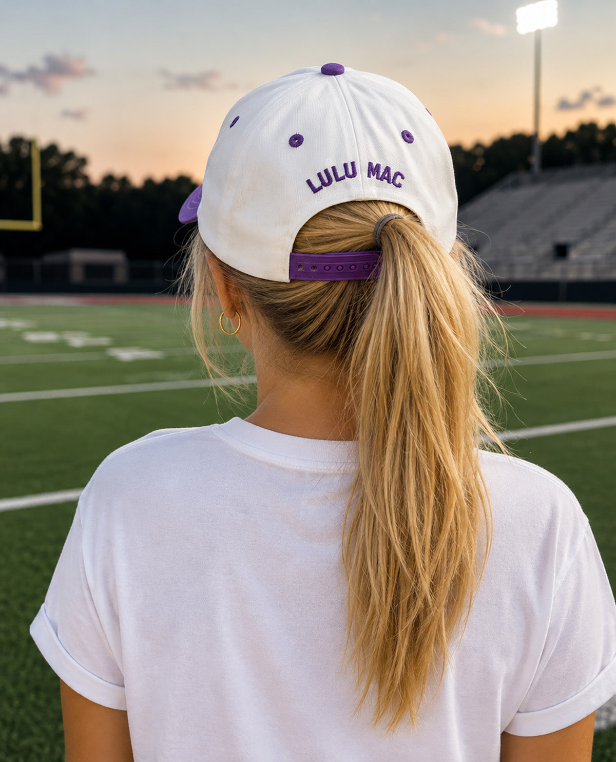 A blonde woman seen from behind wearing a white baseball cap with purple accents and 'LULU MAC' embroidered on the back, standing on a football field at sunset.