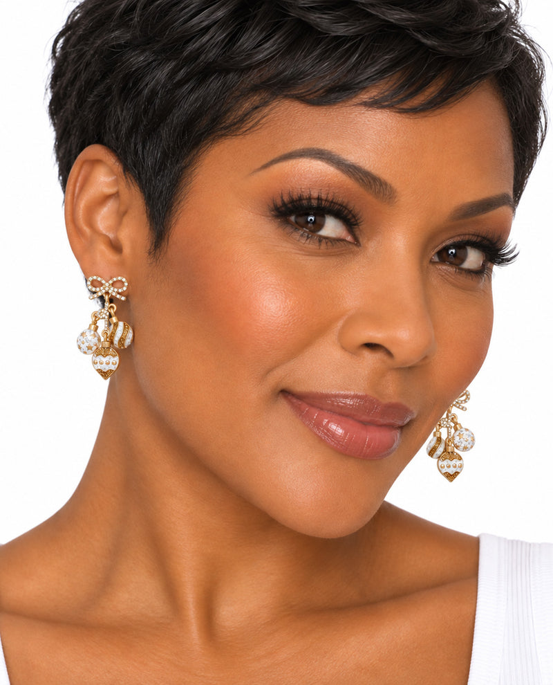 A close-up portrait of a woman with short dark hair wearing ornate gold and white dangling earrings against a plain white background.