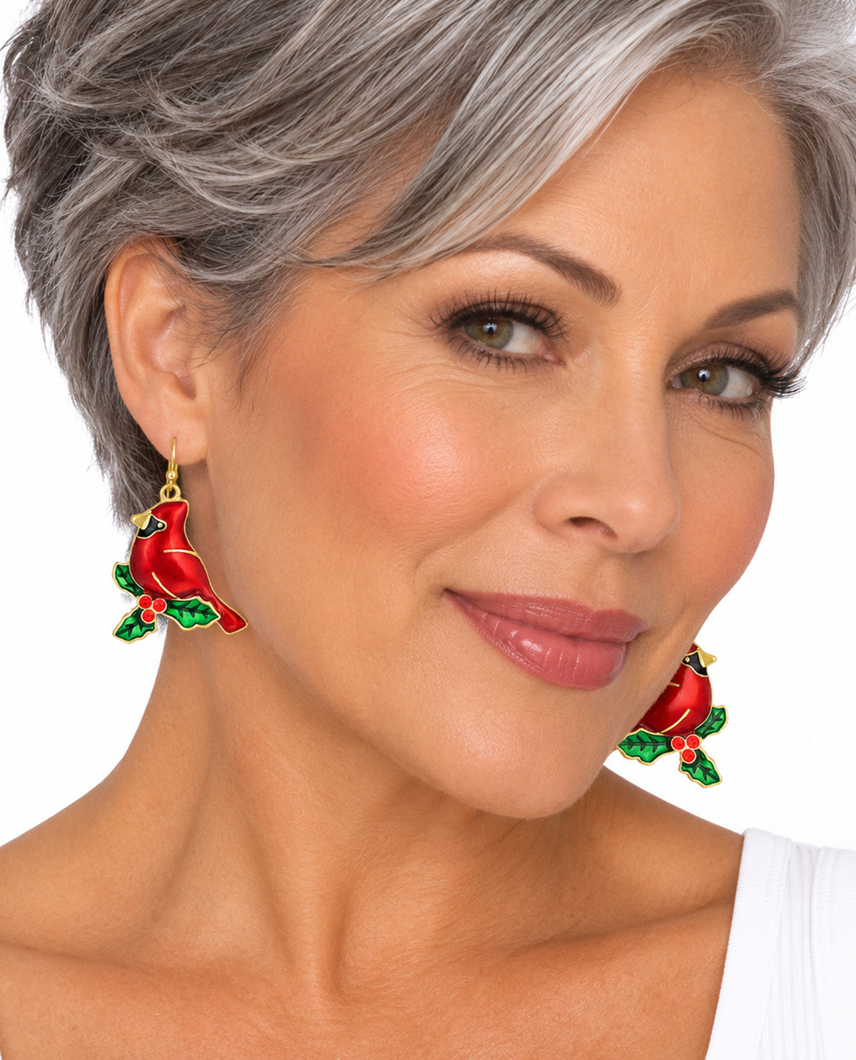A close-up of a woman with short gray hair wearing red and green bird-shaped drop earrings against a plain white background.