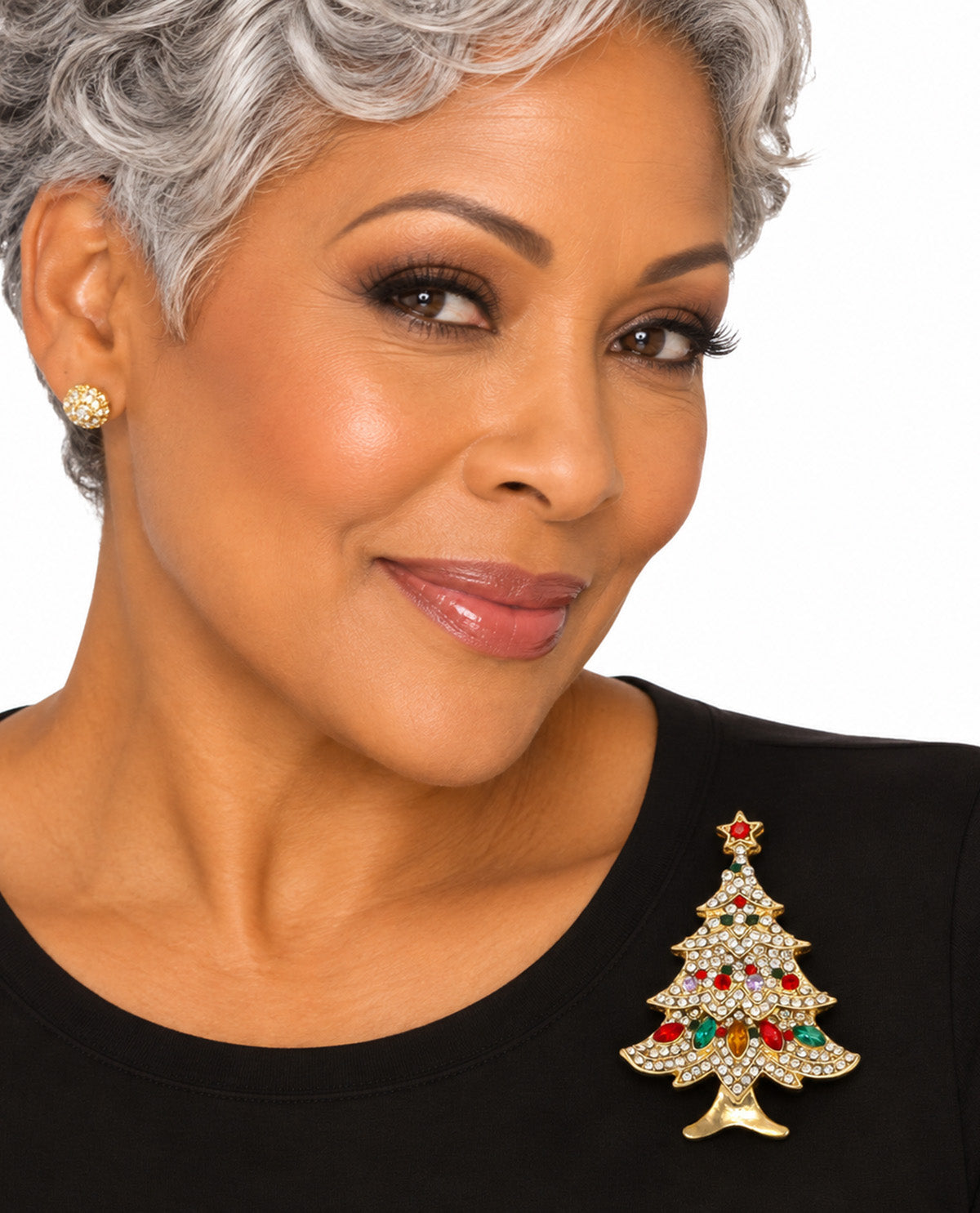 A close-up portrait of a woman with short gray hair wearing a gold Christmas tree brooch embellished with clear rhinestones and red and green crystal accents, paired with a gold stud earring and a black top against a white background.