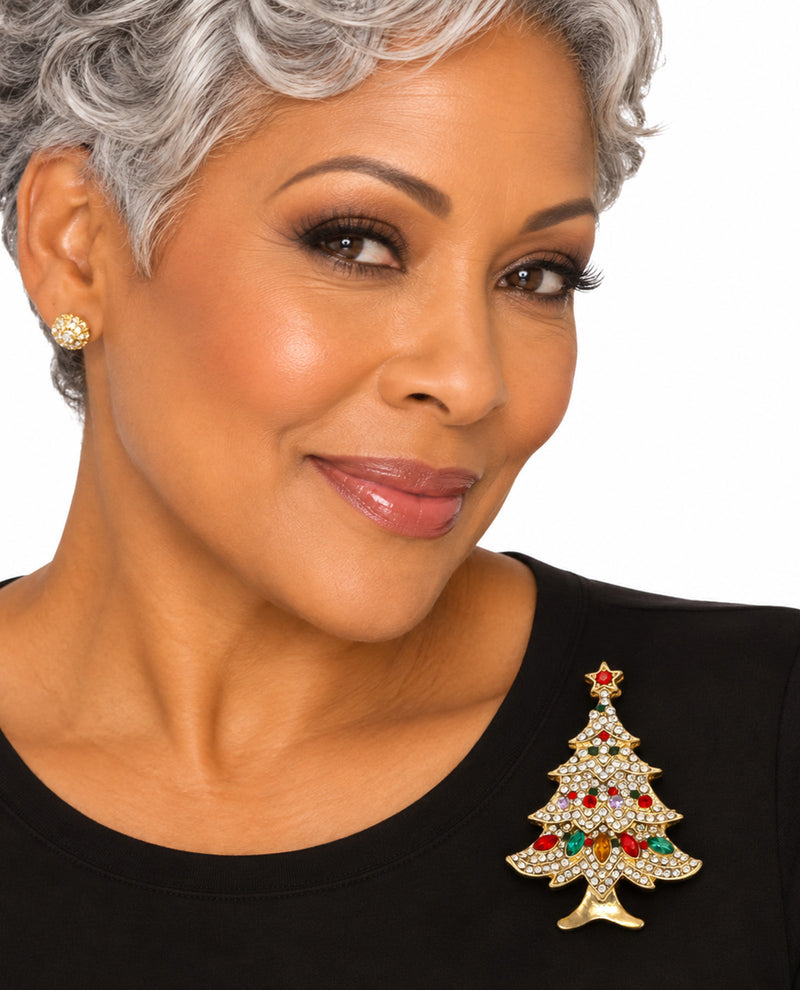 A close-up portrait of a woman with short gray hair wearing a gold Christmas tree brooch embellished with clear rhinestones and red and green crystal accents, paired with a gold stud earring and a black top against a white background.