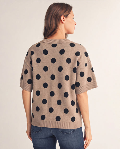 A woman shown from the back wearing a beige knit sweater with large black polka dots and blue jeans, photographed against a plain light beige studio background.