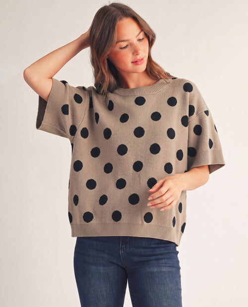 A woman wearing a beige short-sleeve knit sweater with large black polka dots, paired with dark blue jeans, photographed against a plain light background.