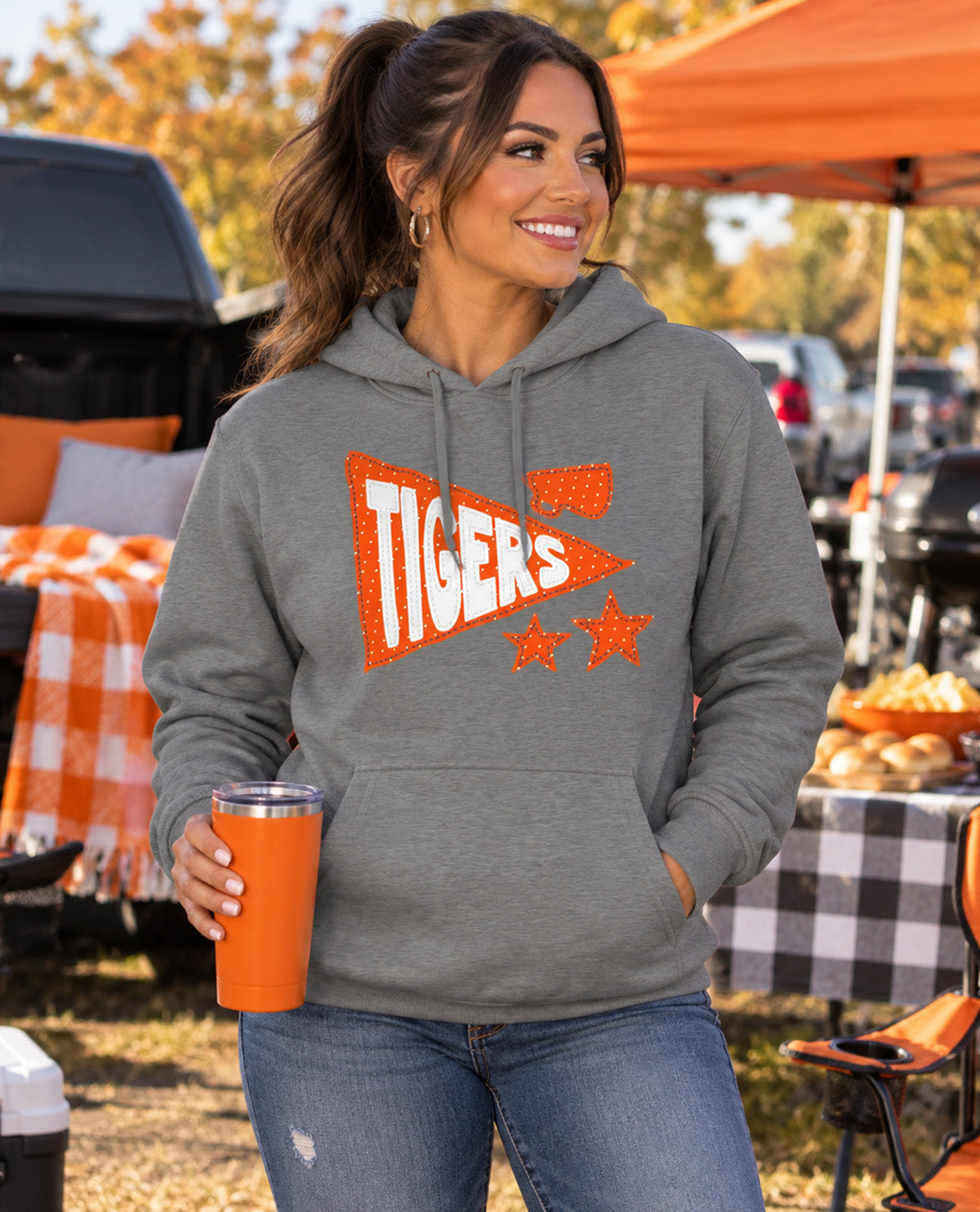 A woman wearing a heather gray hoodie with an orange and white 'Tigers' graphic, holding an orange tumbler at an outdoor autumn picnic.