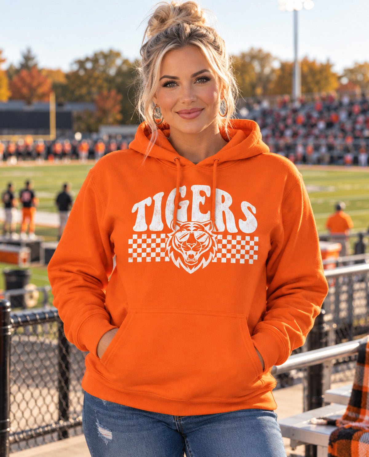 A woman stands on a football field wearing an orange hoodie with a white 'TIGERS' graphic and tiger head logo across the chest, paired with blue jeans.