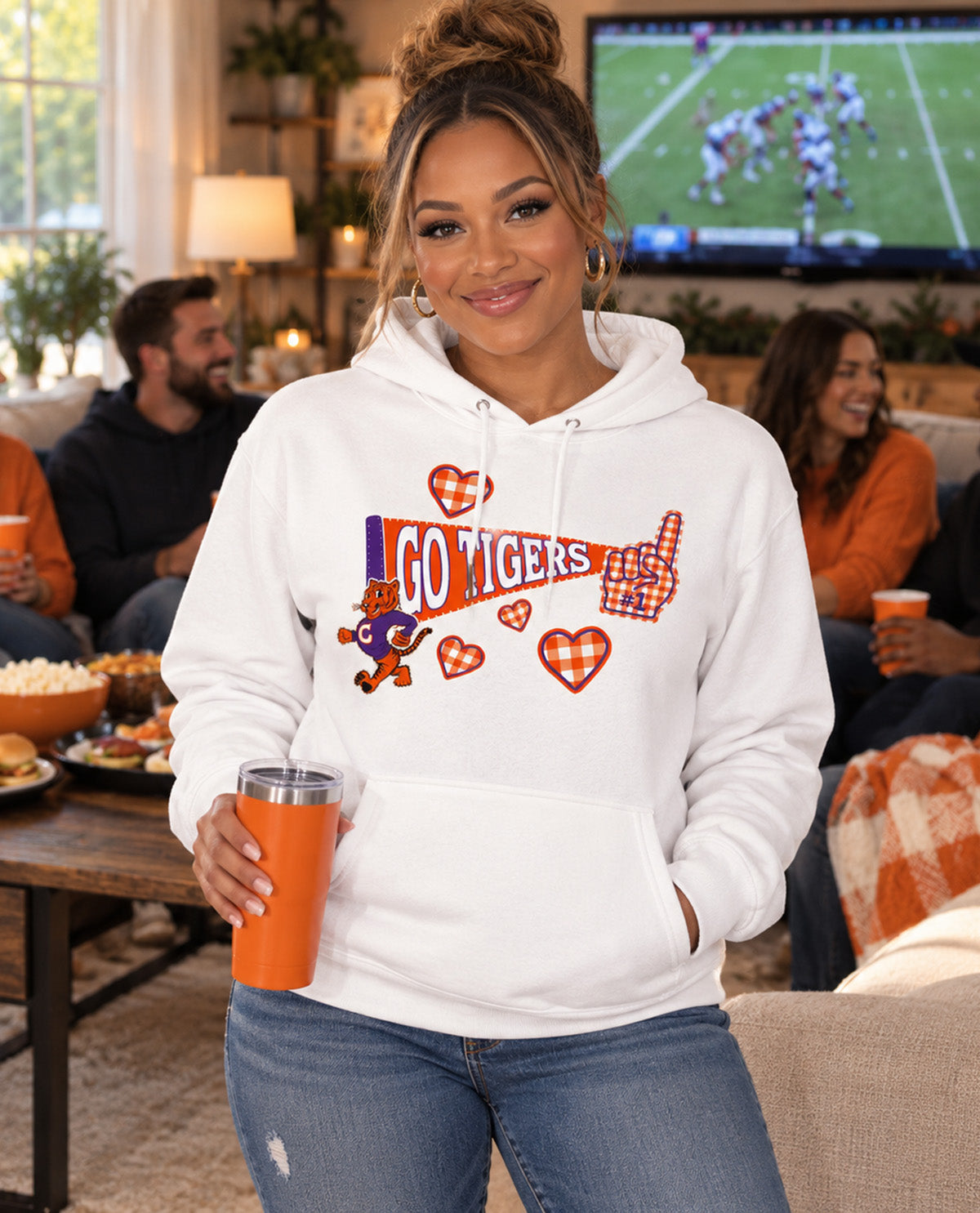 A woman wearing a white hoodie with a 'GO TIGERS' graphic stands in a cozy living room setting, holding an orange tumbler. The hoodie features a graphic with the text 'GO TIGERS', a tiger mascot, and plaid heart details.