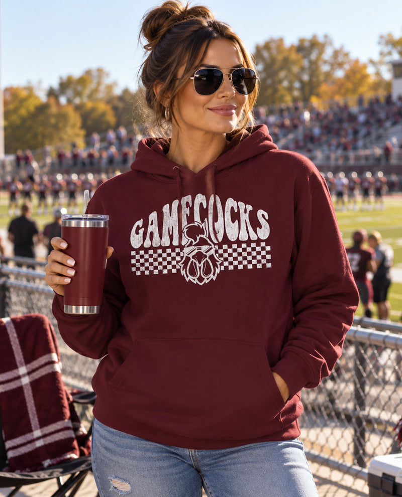 A woman wearing a maroon hoodie with a white 'GAMECOCKS' graphic stands at a sports field, holding a stainless steel tumbler. The hoodie features a white graphic with the word 'GAMECOCKS' in arched block letters, a duck logo, and a checkered stripe design.