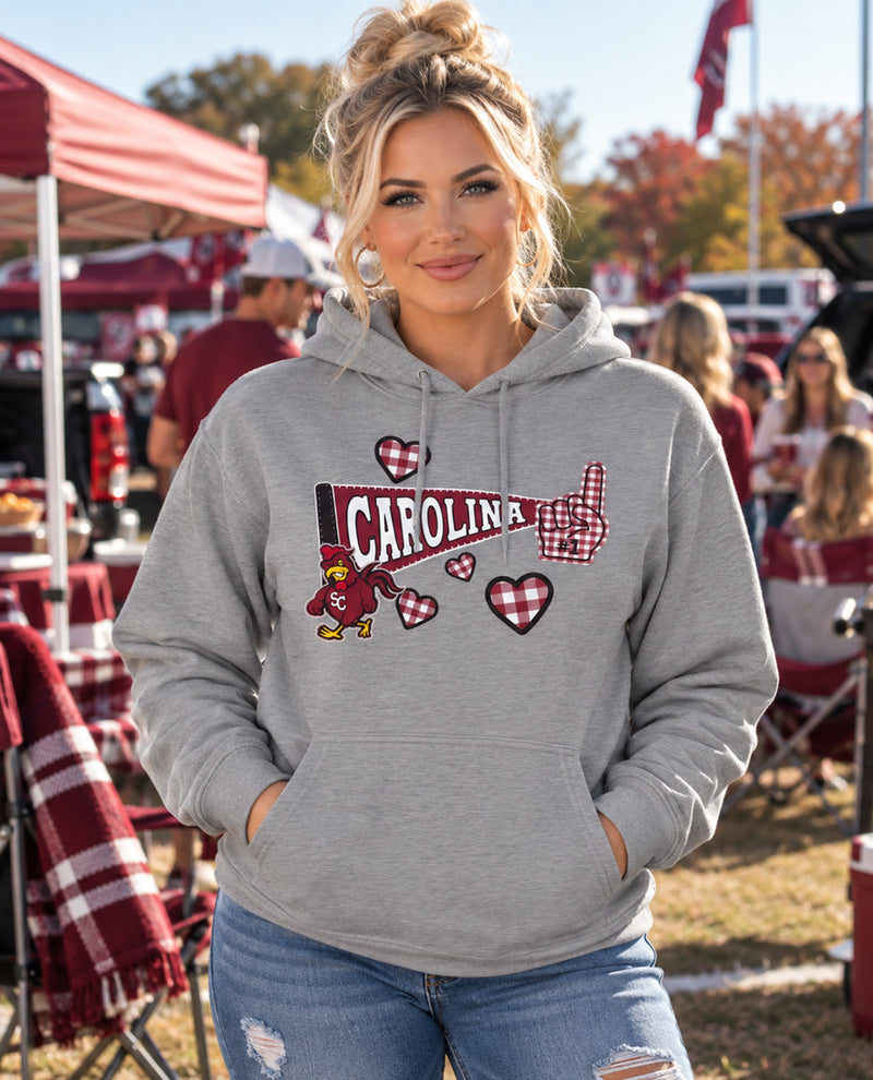 A woman wearing a heather gray hoodie with a 'Carolina' graphic featuring red hearts, a small mascot, and a thumbs-up, photographed outdoors at what looks like a tailgate or event.