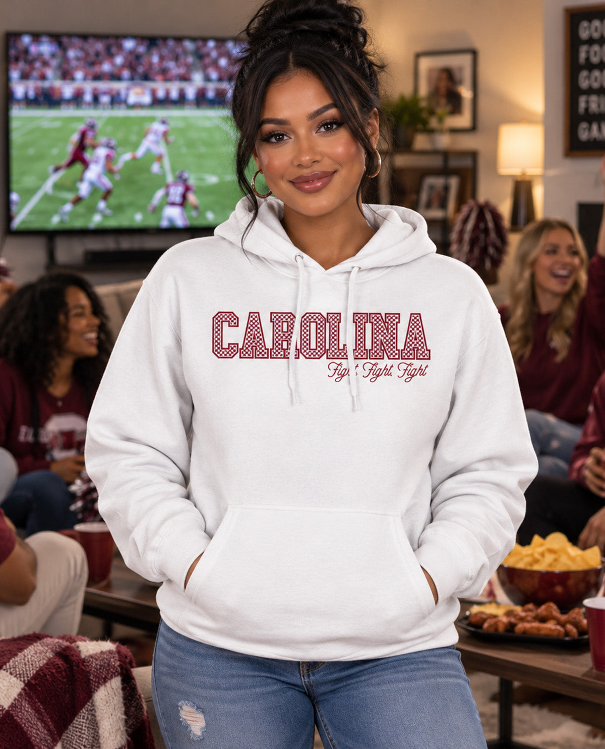 A woman wearing a white pullover hoodie with 'CAROLINA' and 'FIGHT FIGHT FIGHT' printed in maroon across the chest, standing in a cozy living room with a TV in the background.