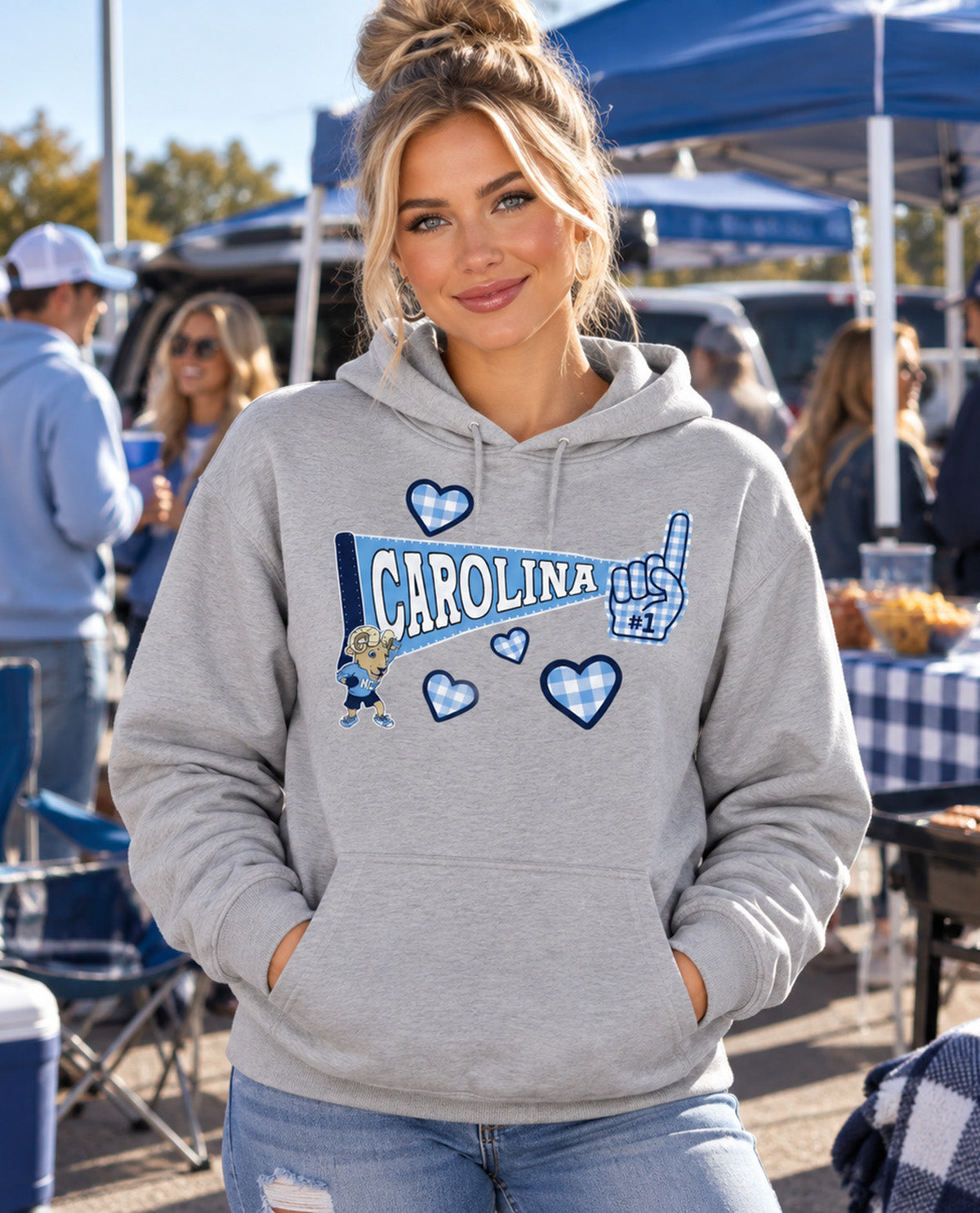 A woman wearing a heather gray hoodie with a blue and white Carolina-themed graphic on the front, standing outdoors at what looks like a tailgate or market event.
