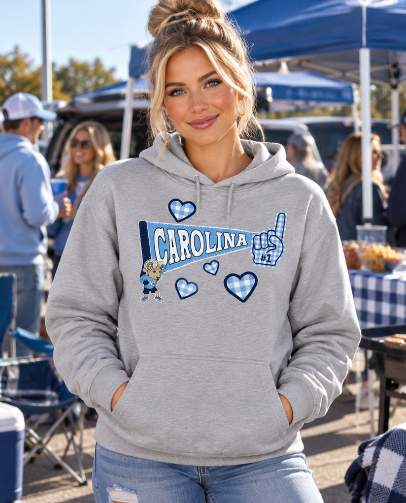 A woman wearing a heather gray hoodie with a blue and white Carolina-themed graphic on the front, standing outdoors at what looks like a tailgate or market event.